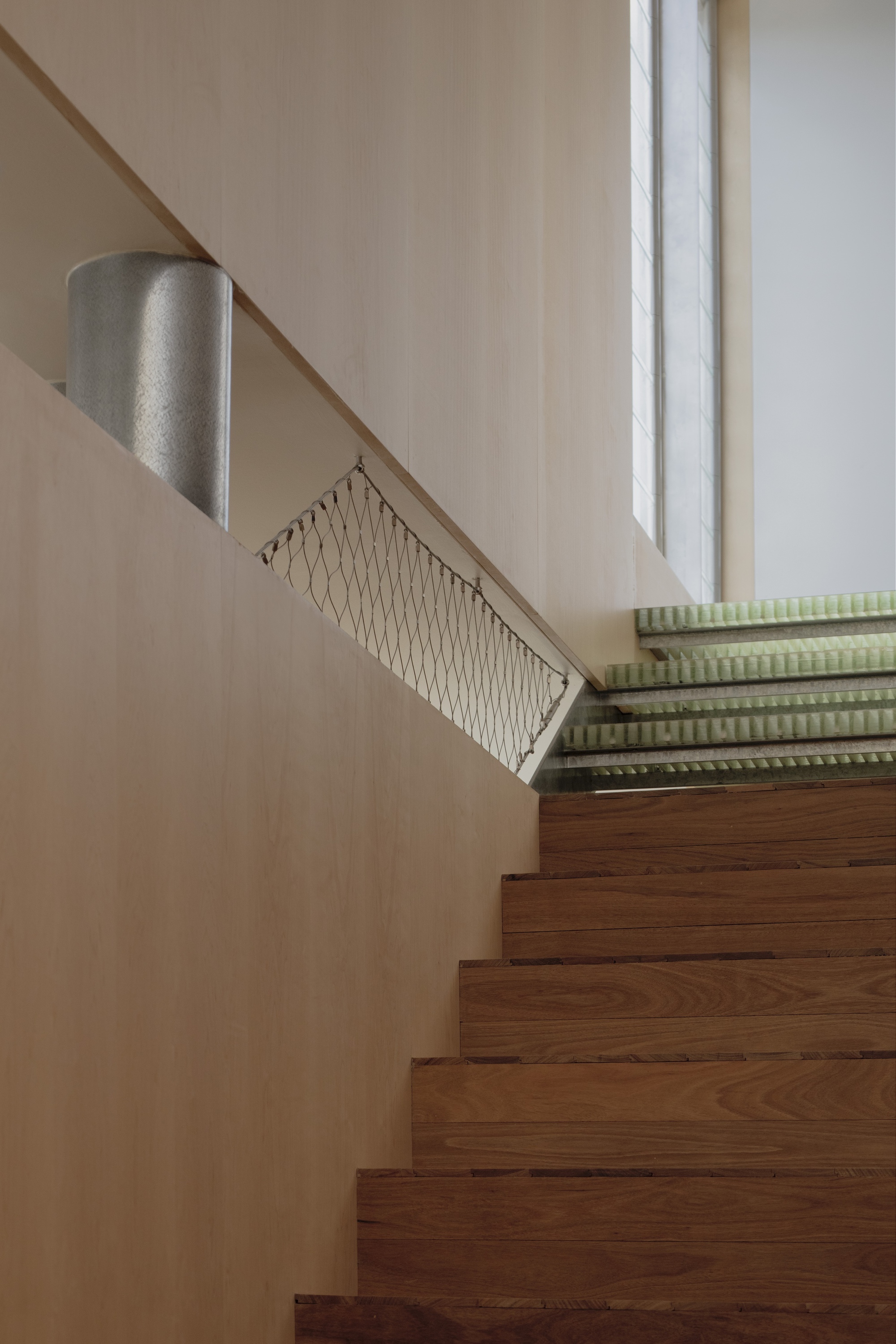 Timber stair rising past a steel mesh balustrade and a metal cylinder toward a tall window with green grating above