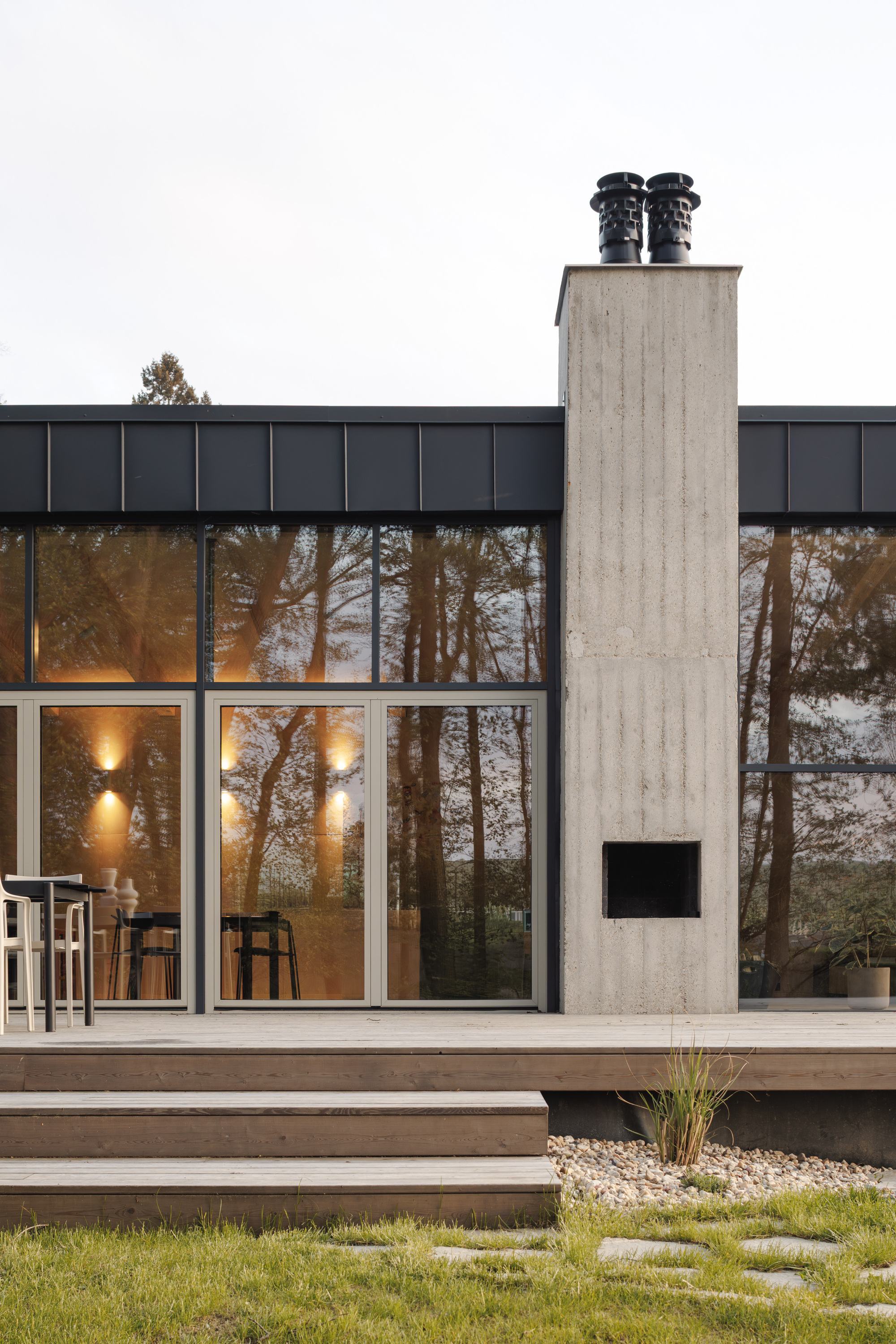 Board-marked concrete chimney with twin black flue caps beside glazed doors, timber steps and pebble bed at dusk