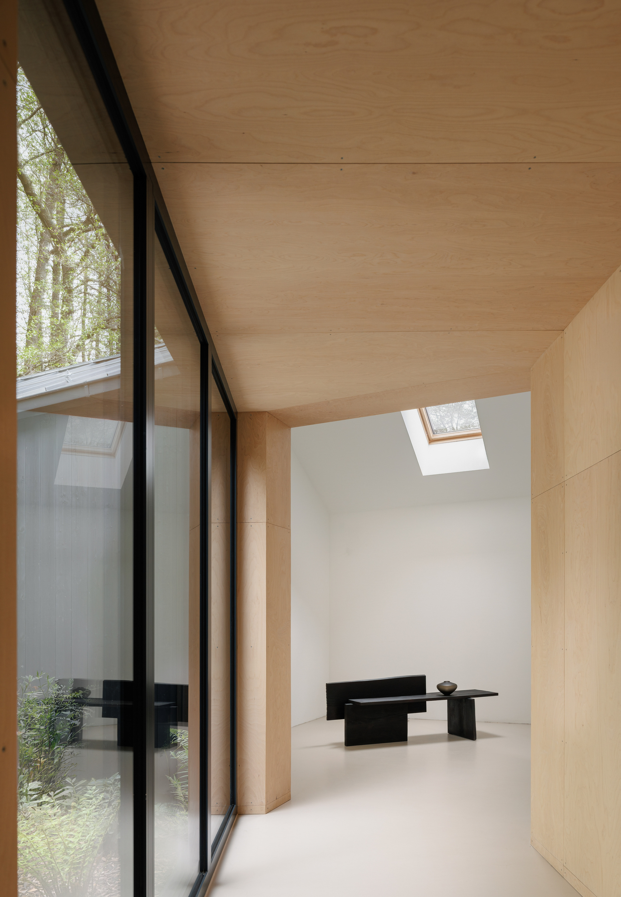 Plywood-lined corridor beside a black-framed glass wall, leading to a white room with a skylight and a dark bench