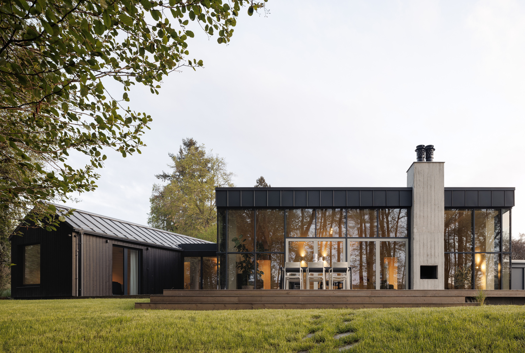 Long facade of dark glazing, a concrete chimney and a black timber outbuilding glowing at dusk behind a lawn