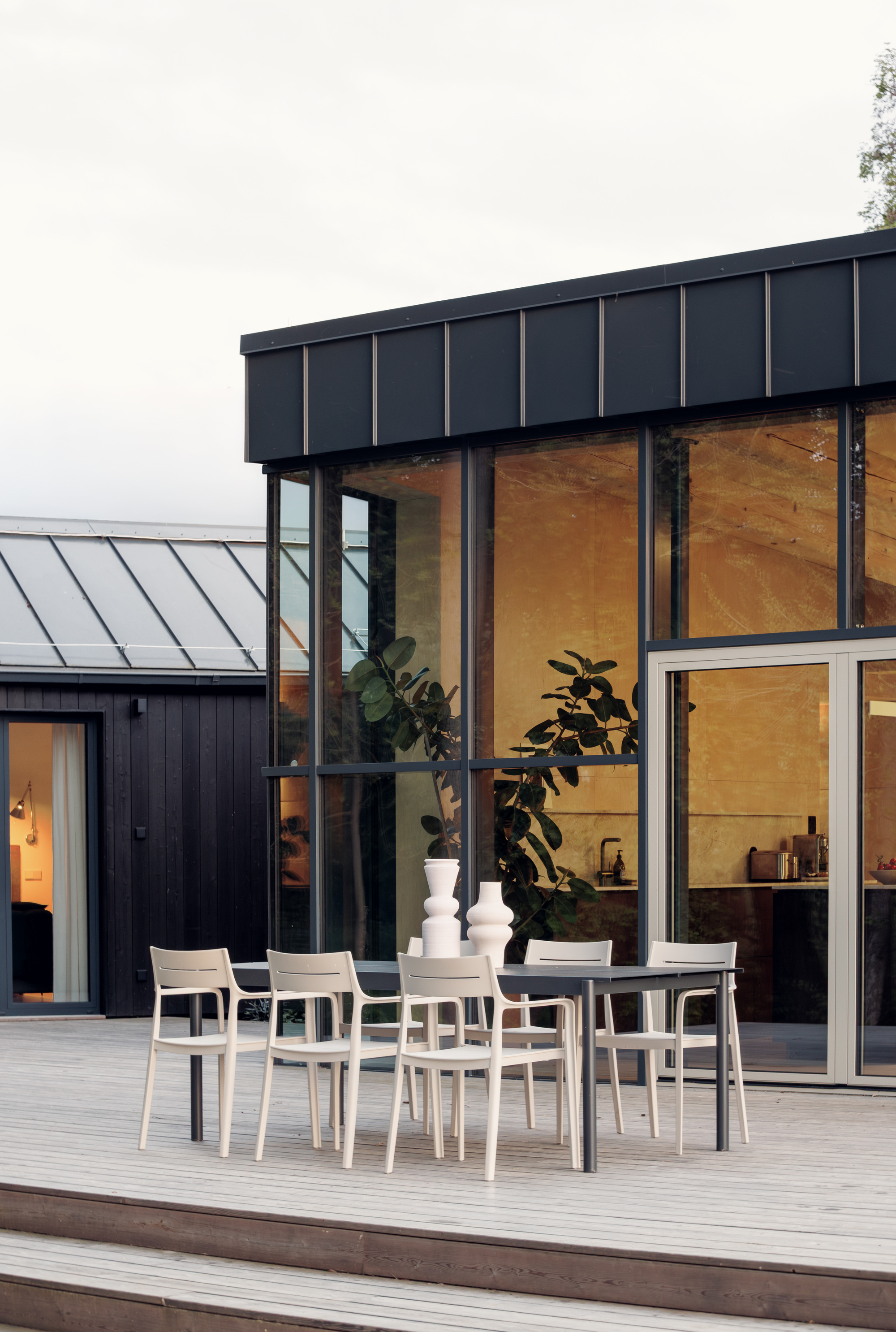 Pale dining chairs and a table on the deck in front of the dark-framed glass corner at dusk