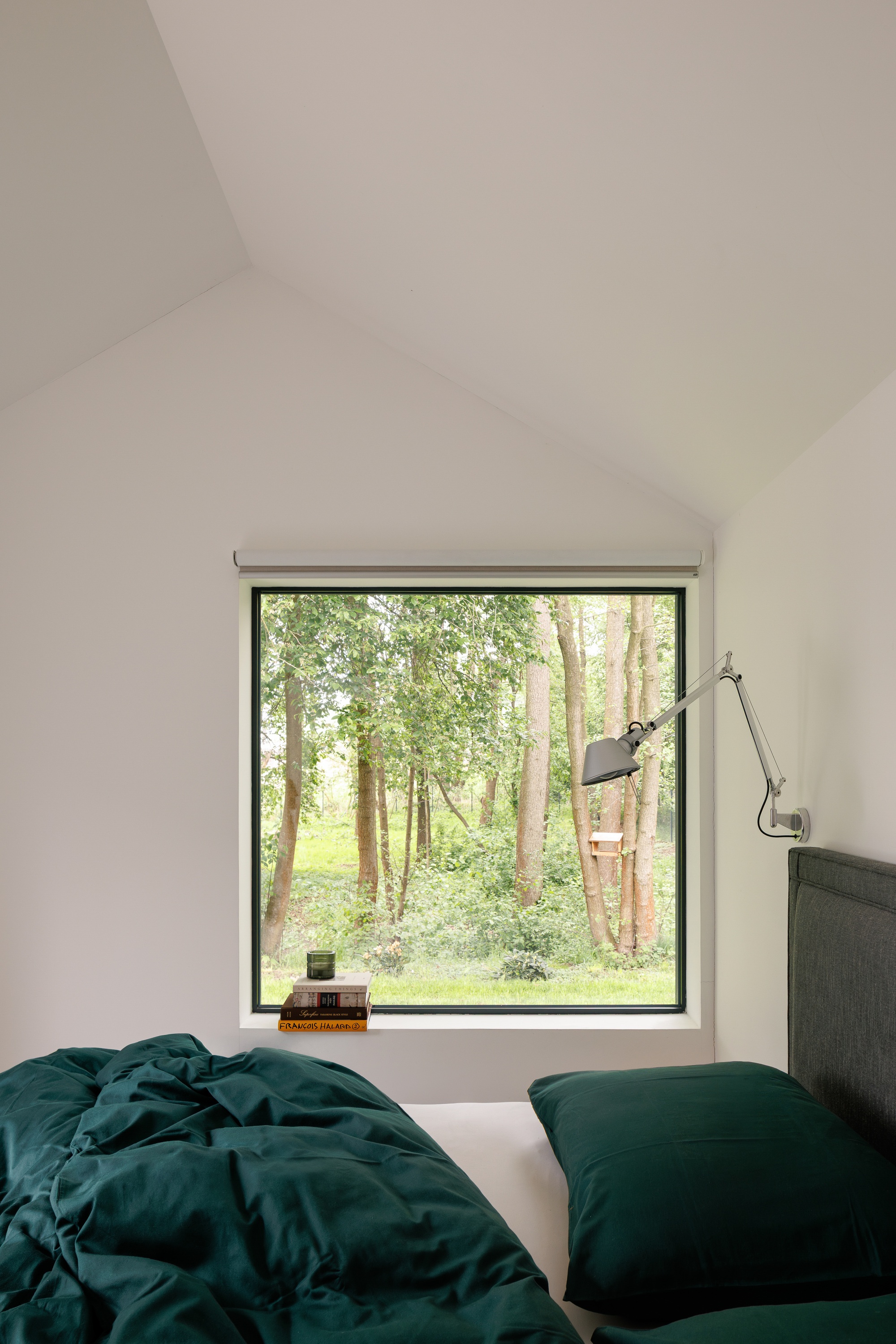 Bedroom with a square window framing birch trees, dark green bedding and an adjustable wall lamp beneath the pitched ceiling