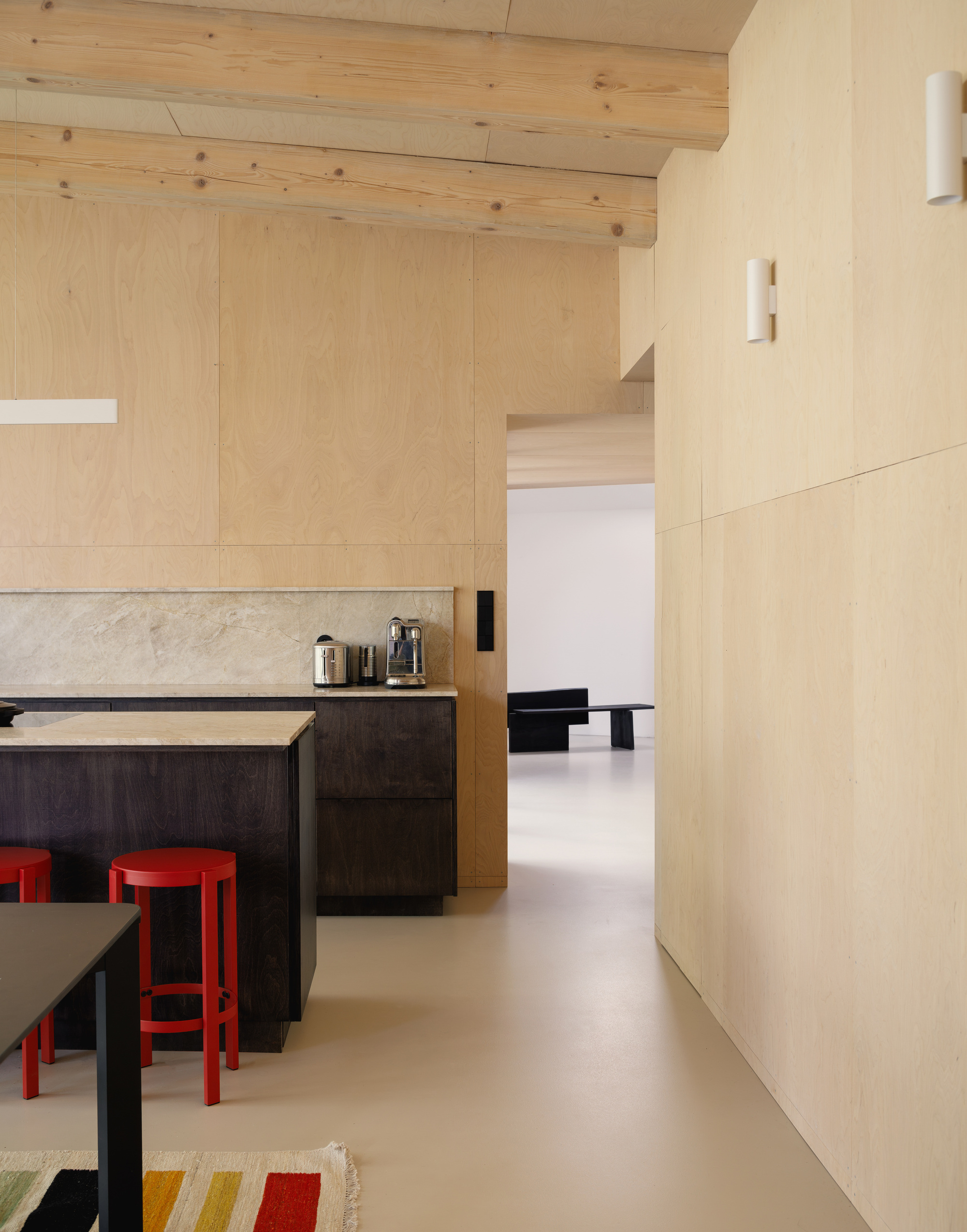 Plywood-clad kitchen with exposed log beams, a dark island with stone top and red stools in soft daylight