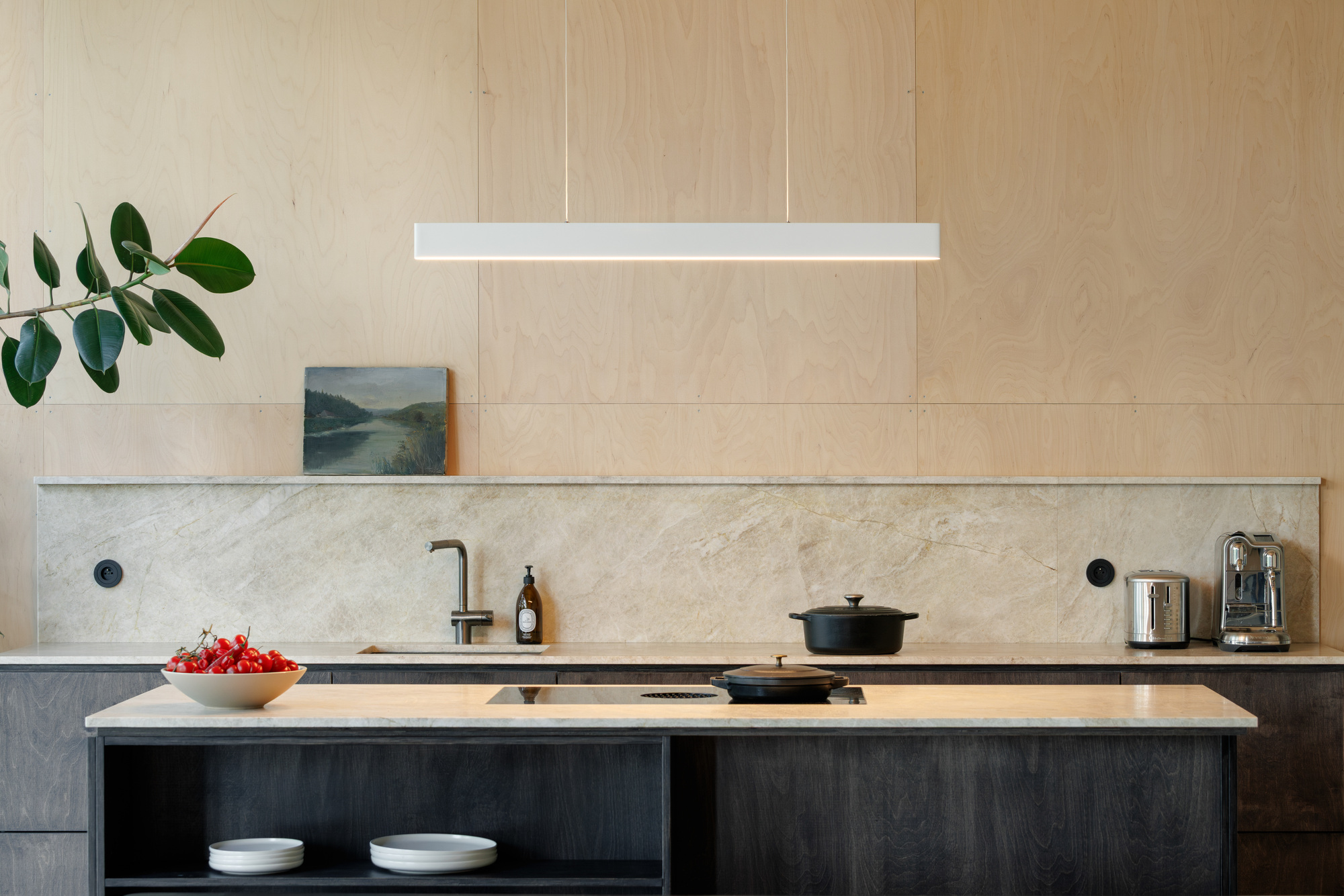 Kitchen island and stone backsplash against a plywood wall, with a linear pendant light and a bowl of red cherries