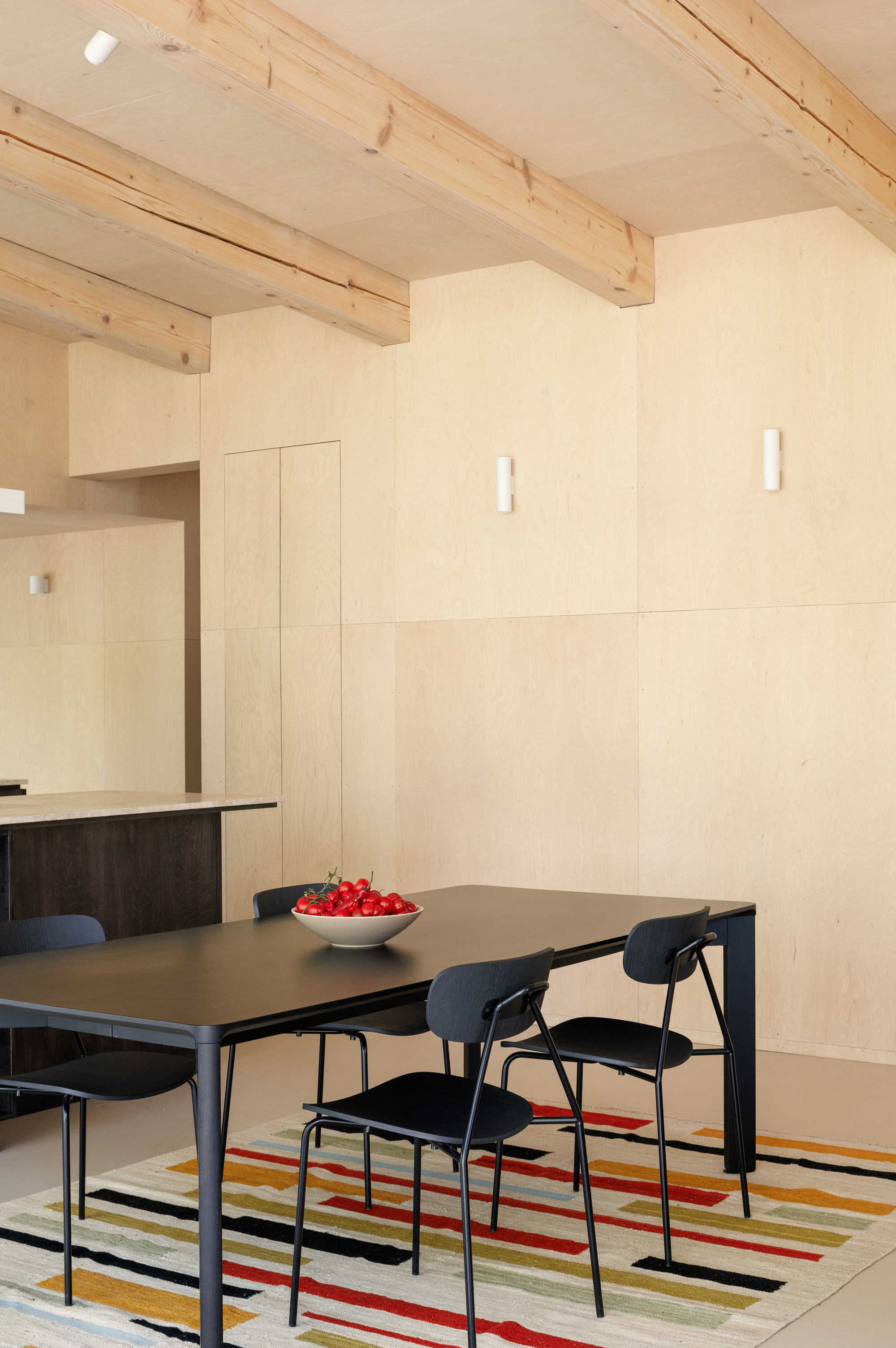 Black dining table and chairs on a striped rug in front of plywood wall panels and exposed timber beams