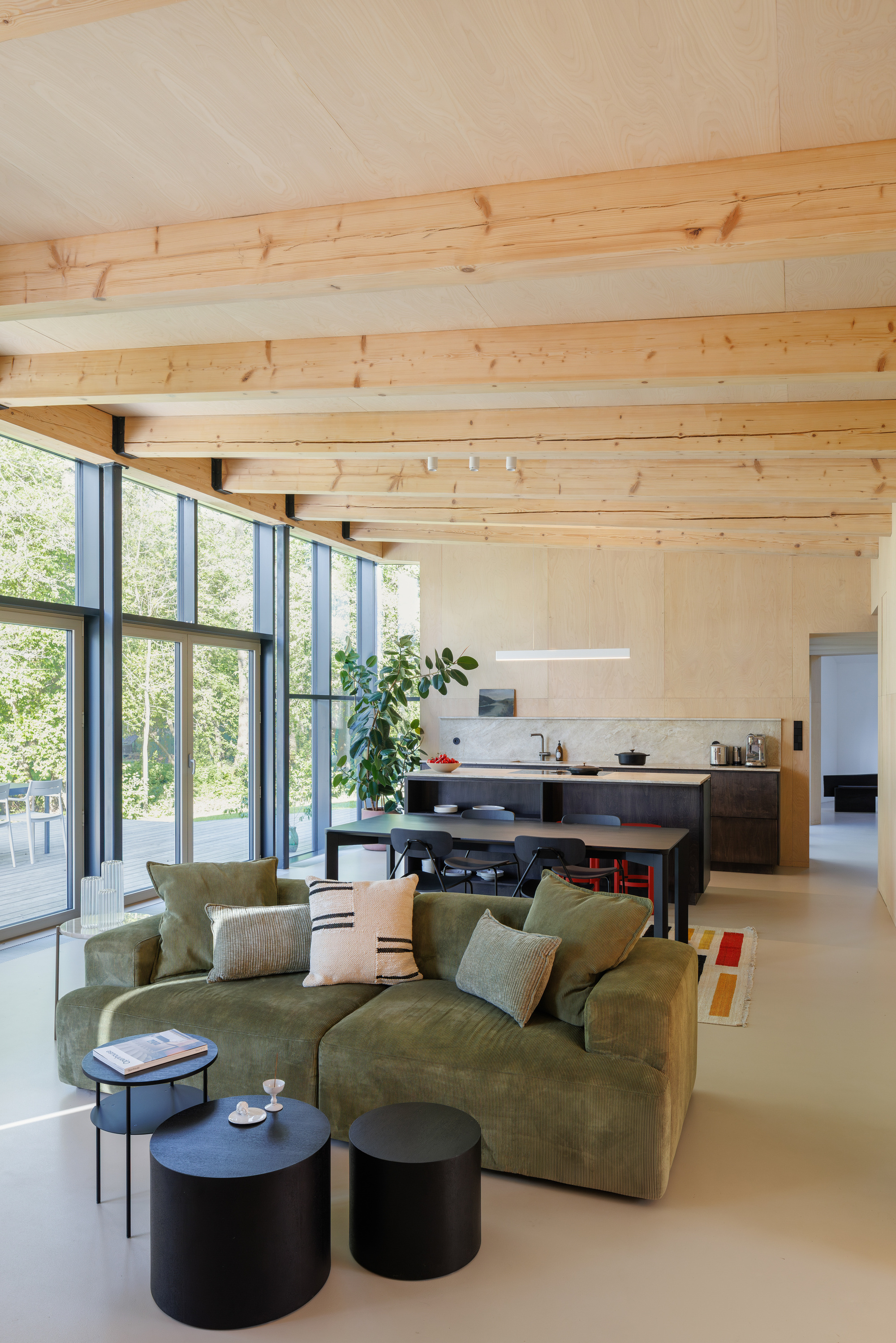 Open living space with a green sofa, black side tables and exposed timber beams, with glazed wall opening to the trees