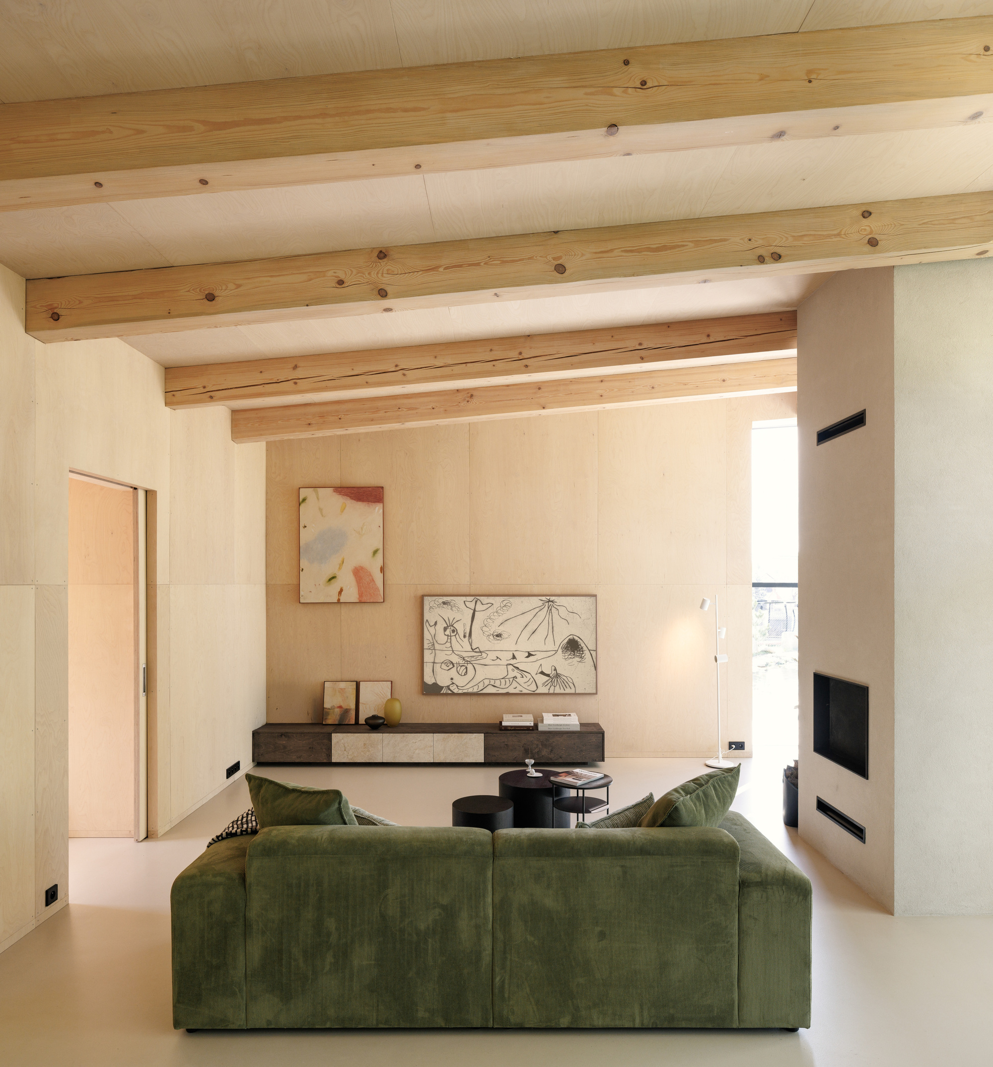 Living room with exposed timber beams, plywood walls and a green velvet sofa facing a recessed fireplace in soft daylight