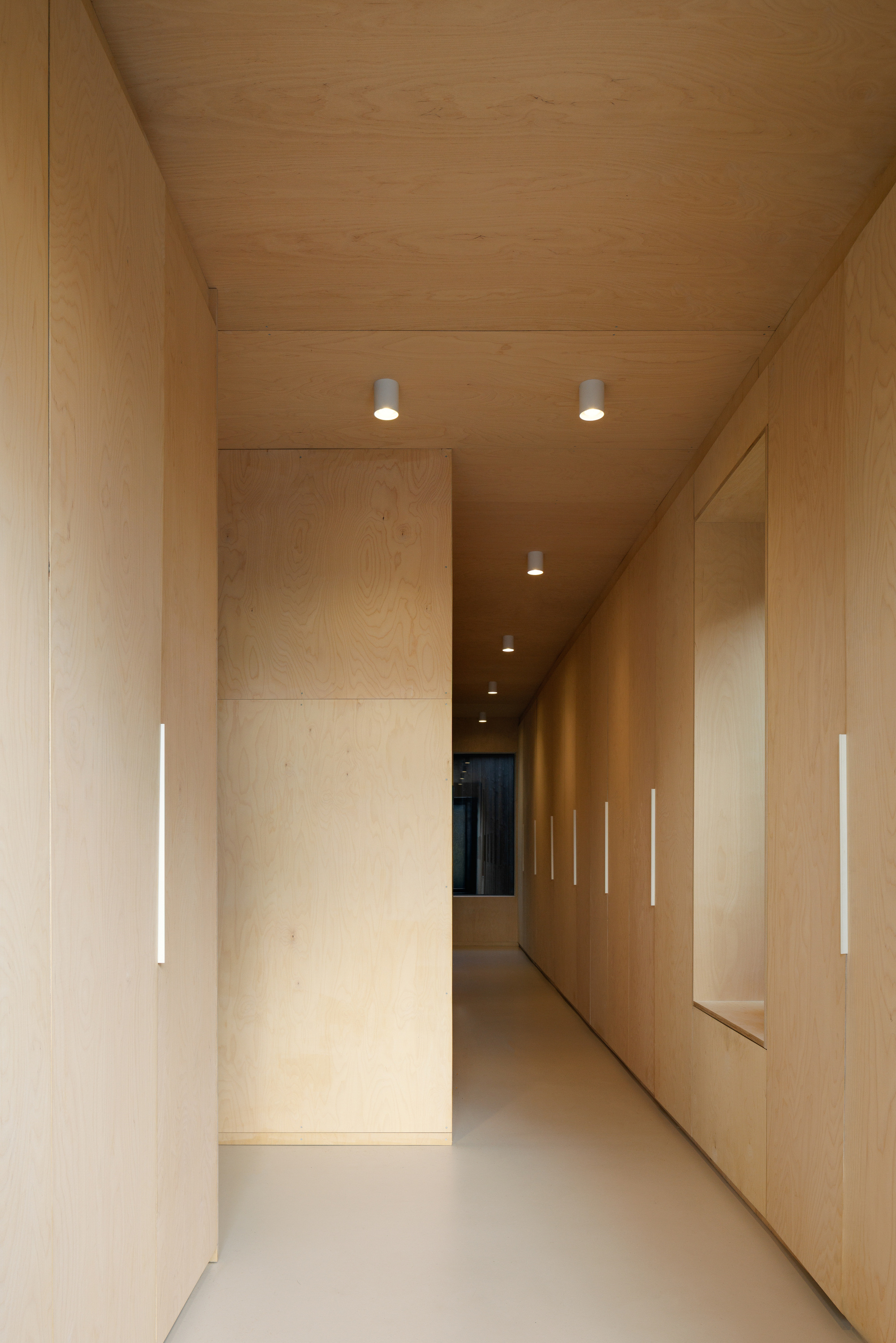 Narrow plywood corridor lined with flush cabinet doors, slim white pulls and a row of ceiling downlights