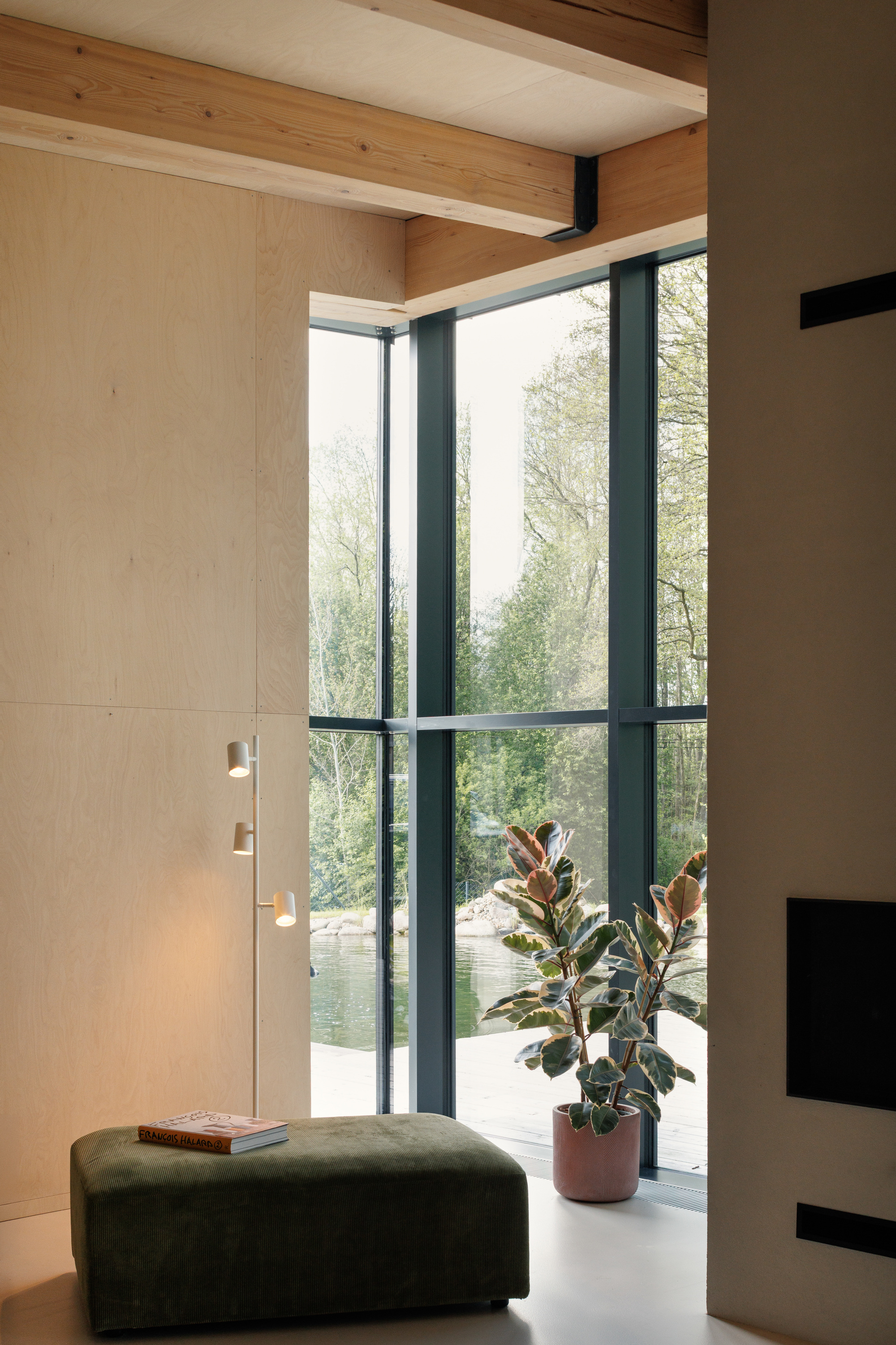 Corner of floor-to-ceiling glazing with a rubber plant, green ottoman and floor lamp, trees and water visible outside in spring