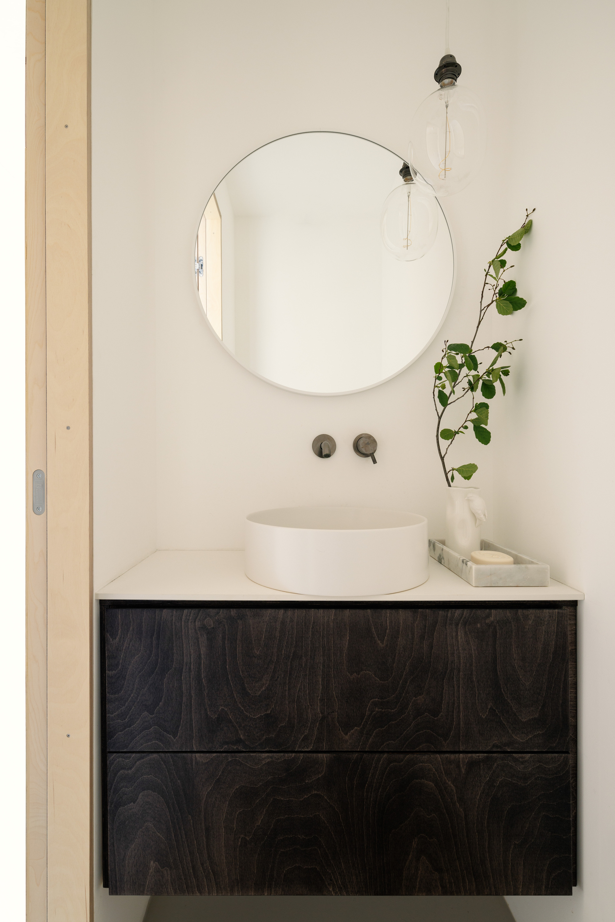 Wall-hung dark stained wood vanity with a round white basin, circular mirror and a leafy branch beside pendant bulbs