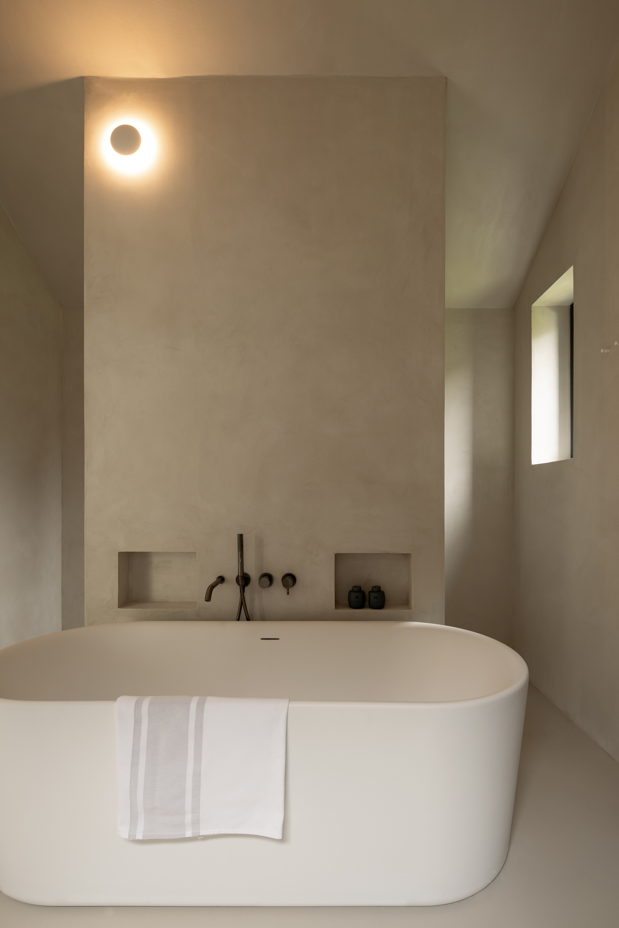 Freestanding white oval bathtub against a tall plastered wall with twin niches, wall-mounted taps and a glowing round sconce