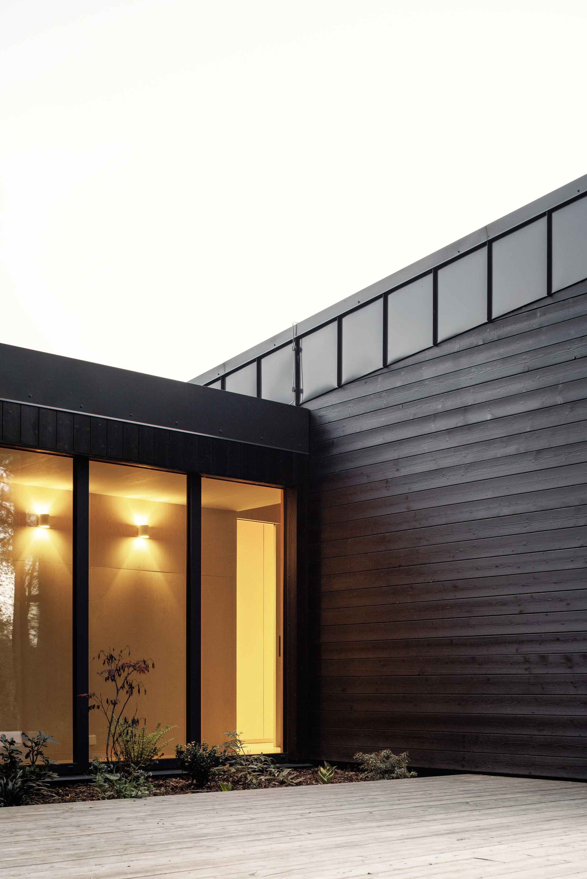 Corner of a black timber-clad volume with glazed entrance, warm wall lights and a deck at dusk