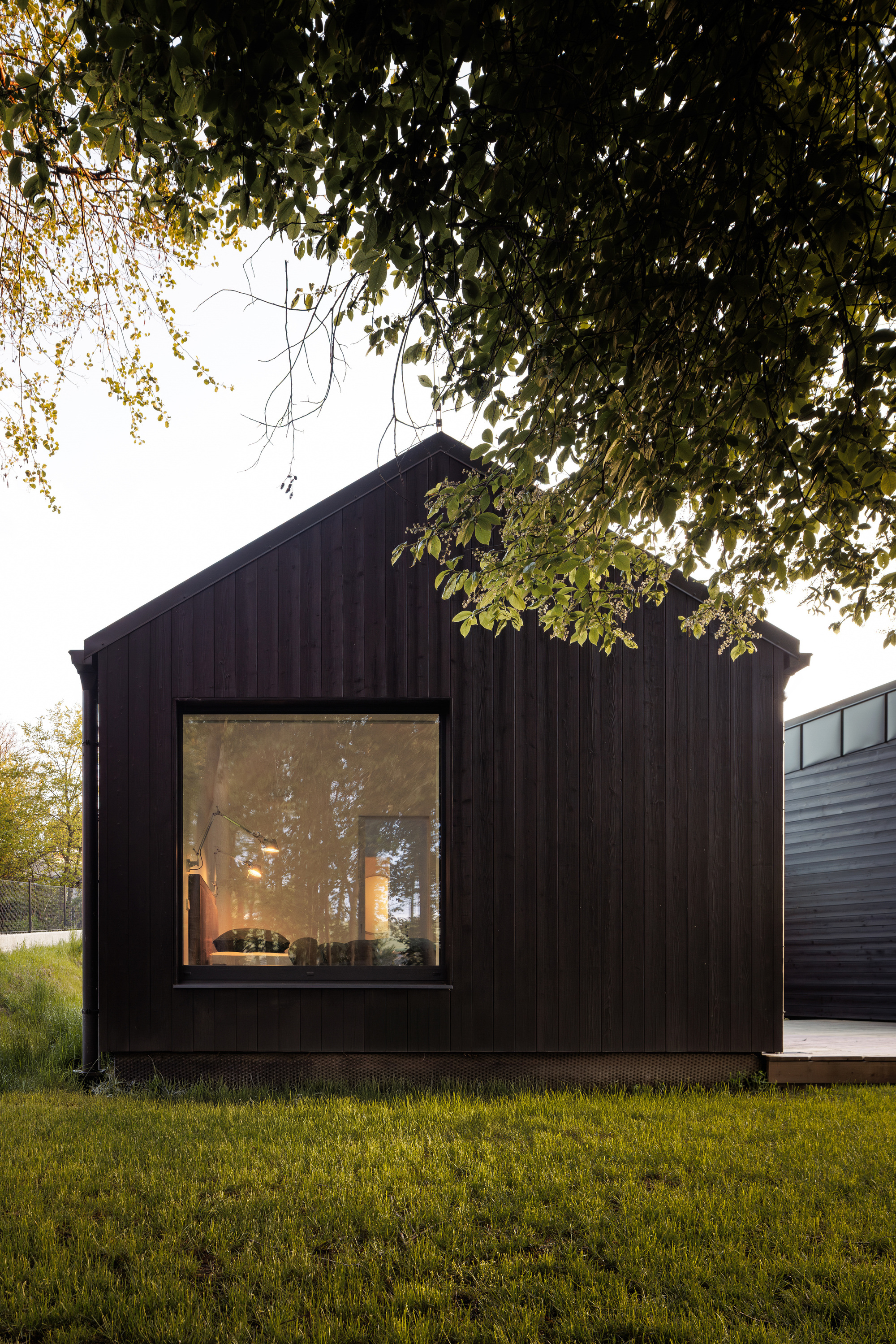 Gable end of a charred timber cabin with a large square window, framed by overhanging leaves and a lawn in soft evening light