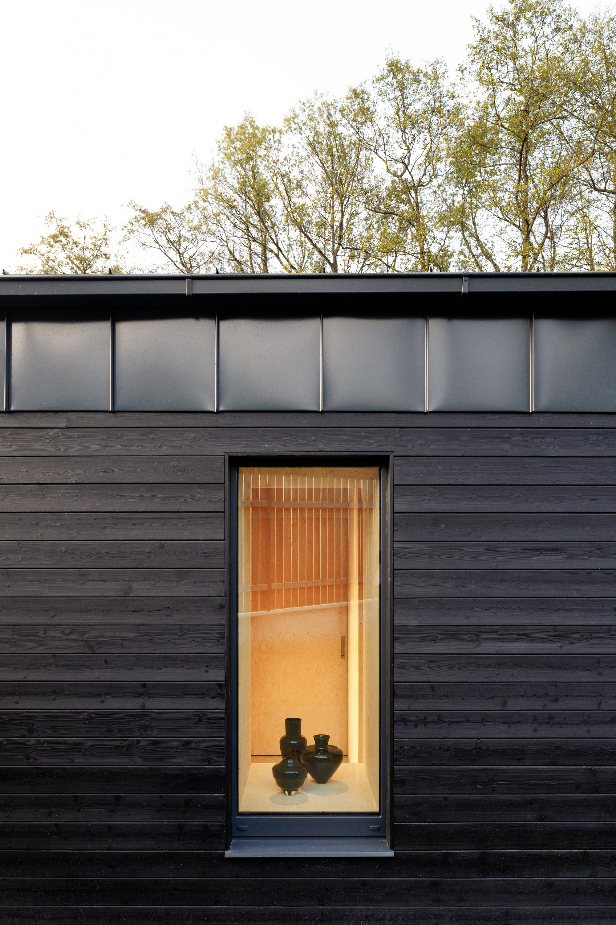 Tall narrow window in dark horizontal timber cladding beneath a standing seam metal fascia, with two black vases lit inside and trees above