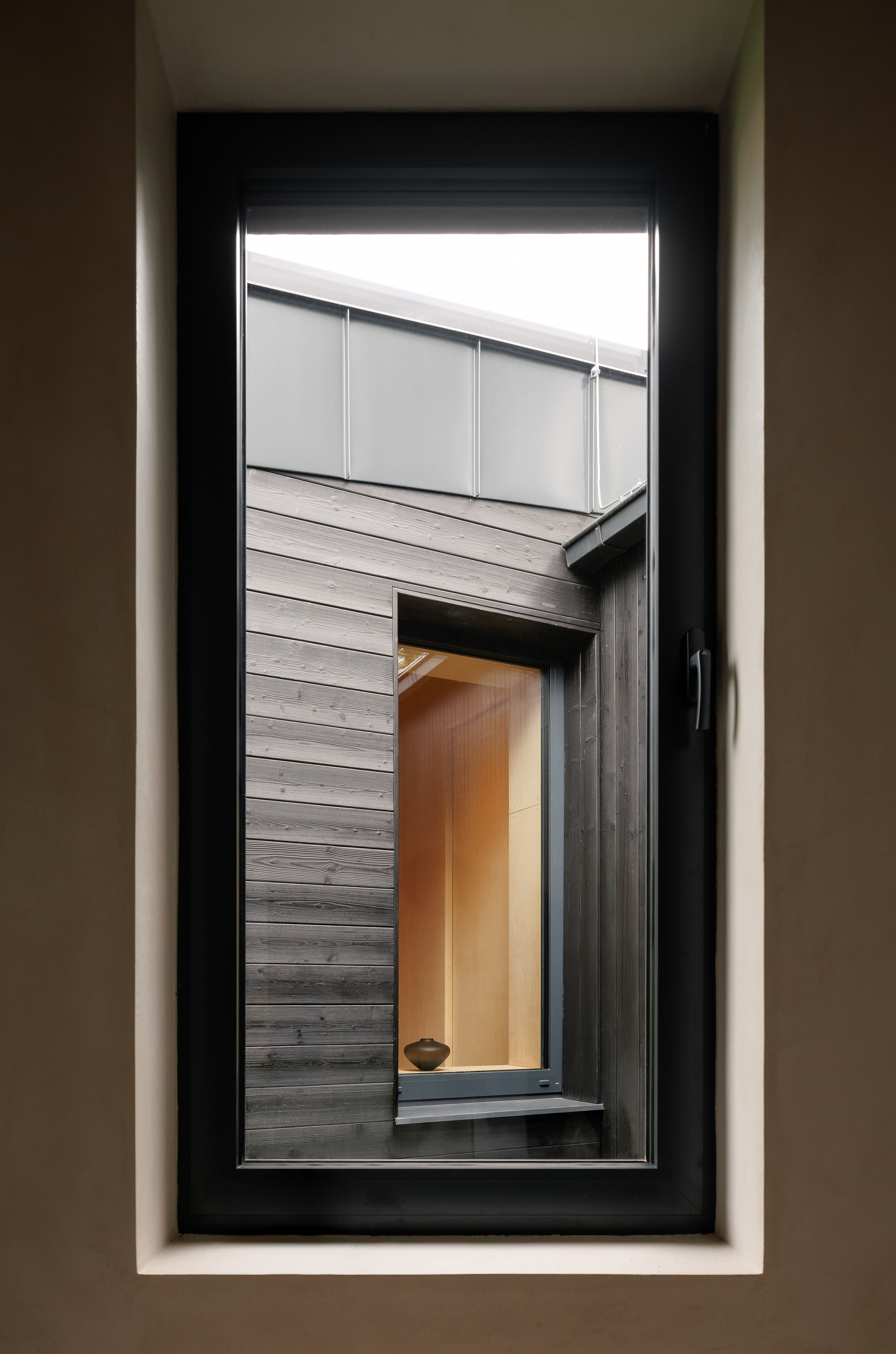 View through a deep-set window reveal toward a neighbouring dark timber wall, metal fascia panels and a glowing wood-lined window