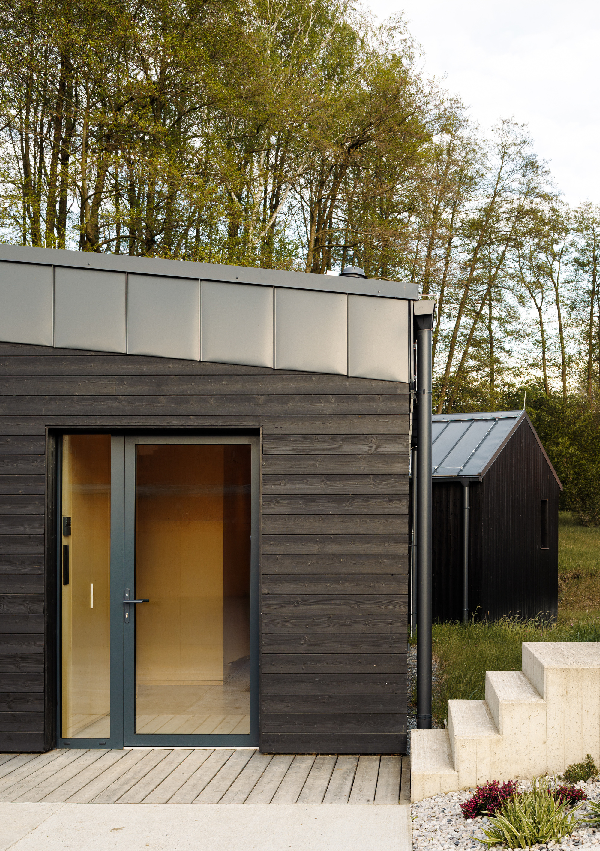 Glazed entry door in dark timber cladding with a metal fascia, a wooden deck, concrete steps and a second cabin among spring trees