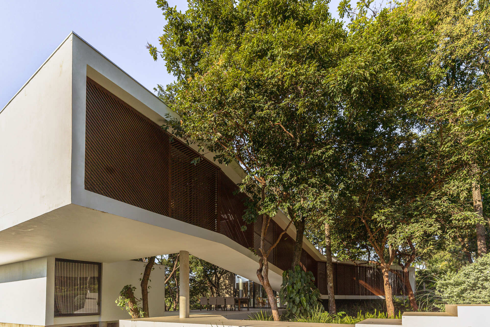 Angled white concrete box with timber louvers cantilevering over a covered terrace and dining chairs, framed by tall trees under clear sky