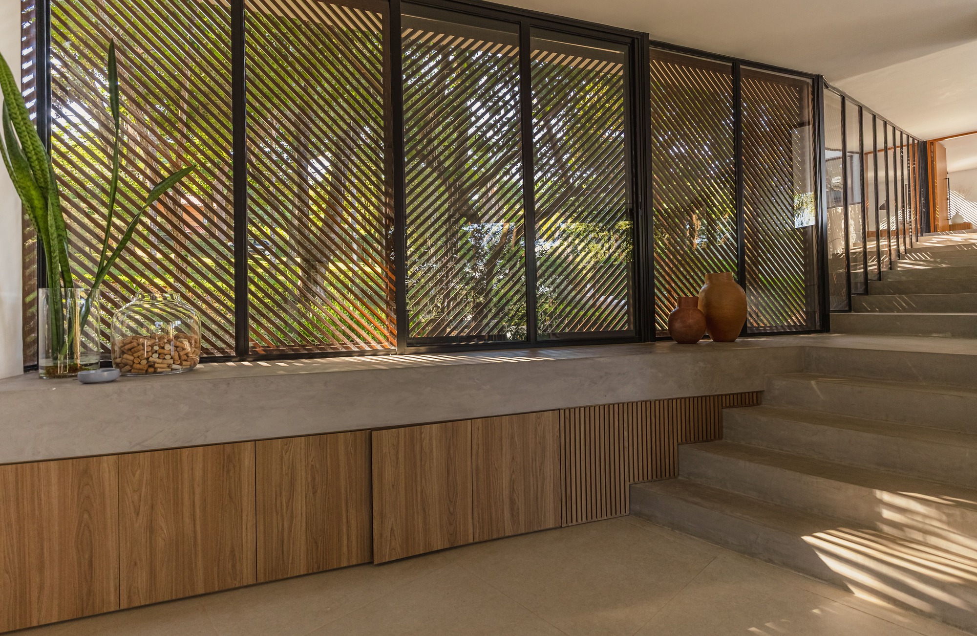 Concrete bench above timber cabinets beside a stair, with diagonal slatted screens filtering the green trees outside