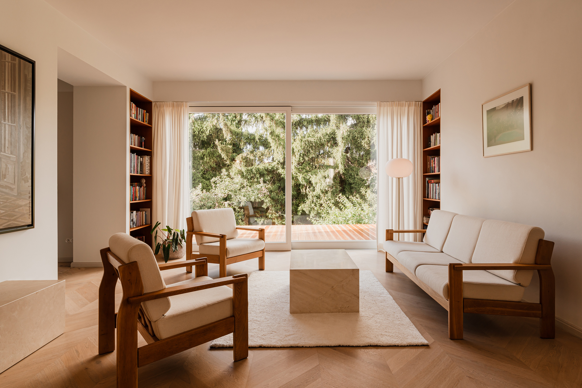 Living room with timber-framed sofa and armchairs on a herringbone floor, facing a sliding glass door onto a deck and spruce trees
