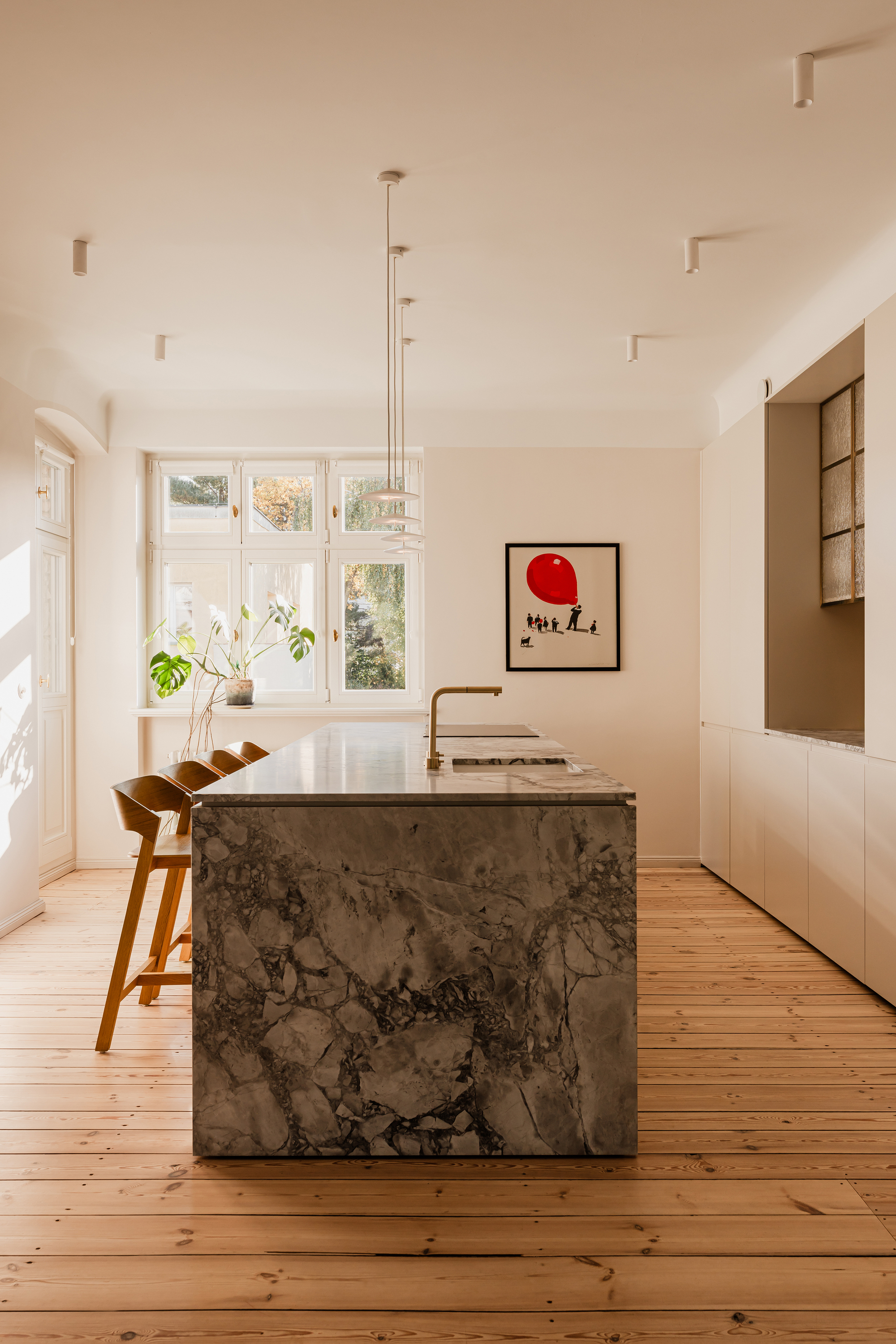 Kitchen island clad in grey veined marble with a brass tap and timber stools, sunlight falling across pine floorboards