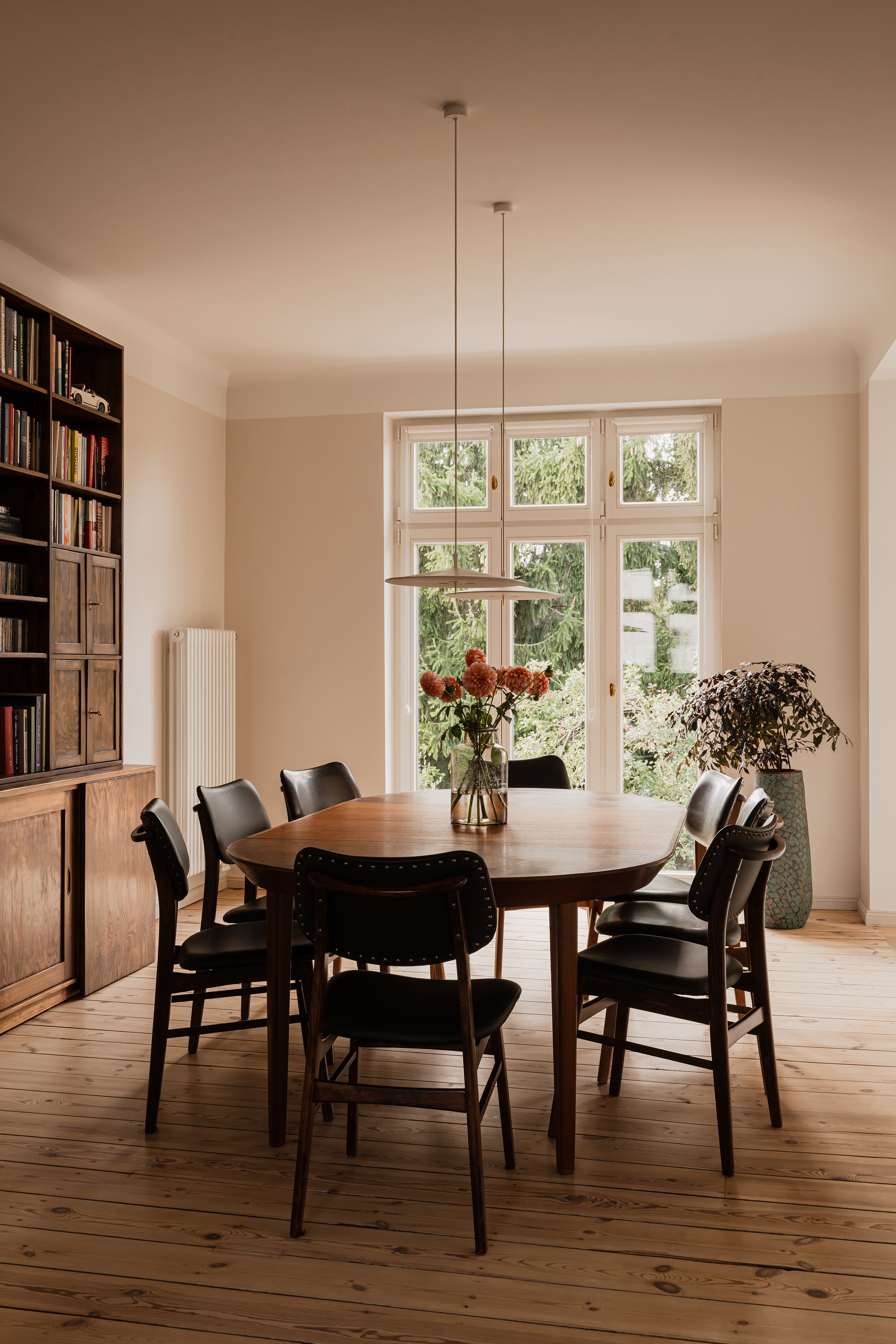 Oval wooden dining table with black leather chairs and a vase of orange flowers before a tall window onto evergreens