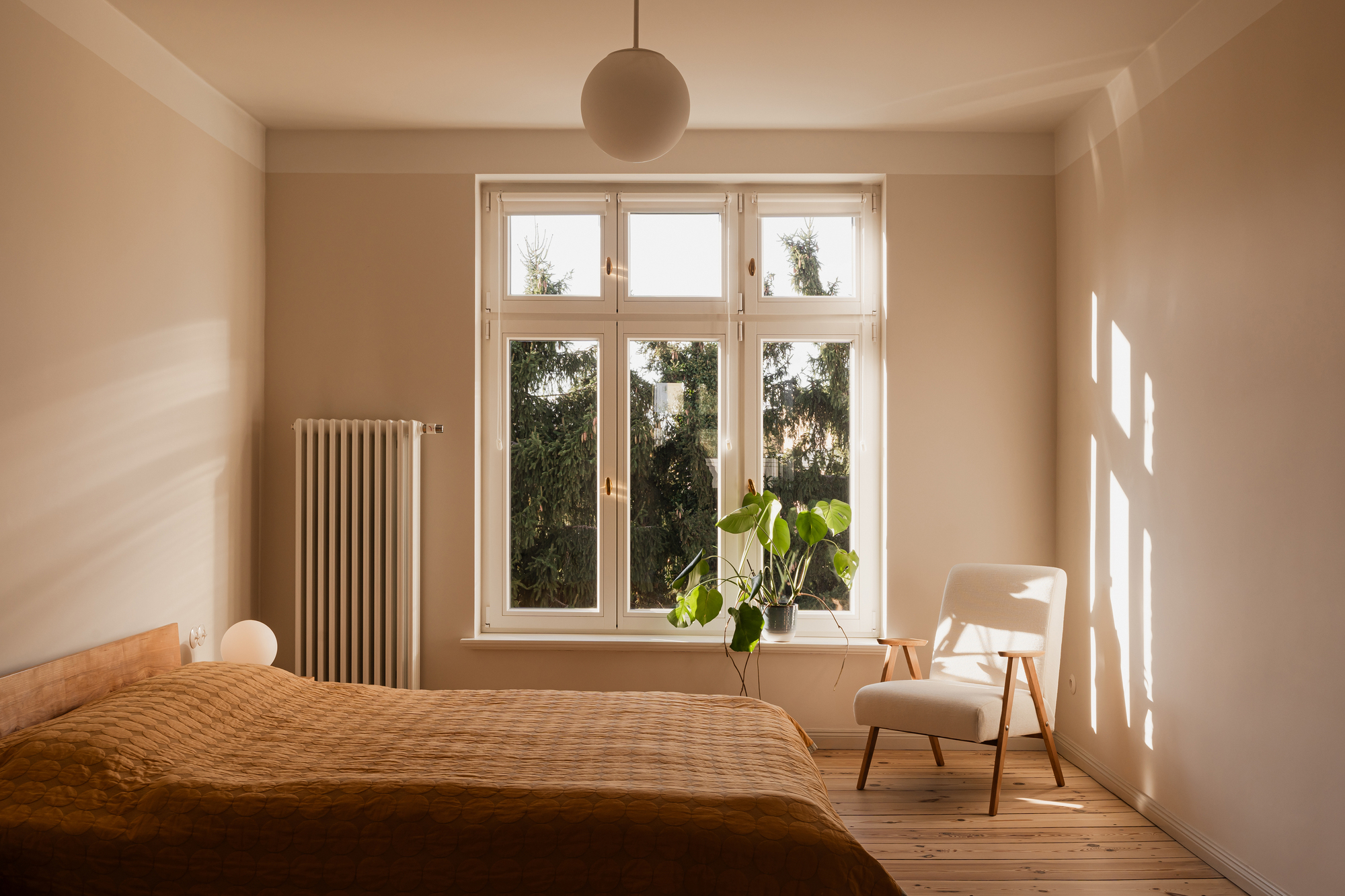 Bedroom with a rust-coloured quilted bedspread and a white armchair, low sun casting window shadows across the wall