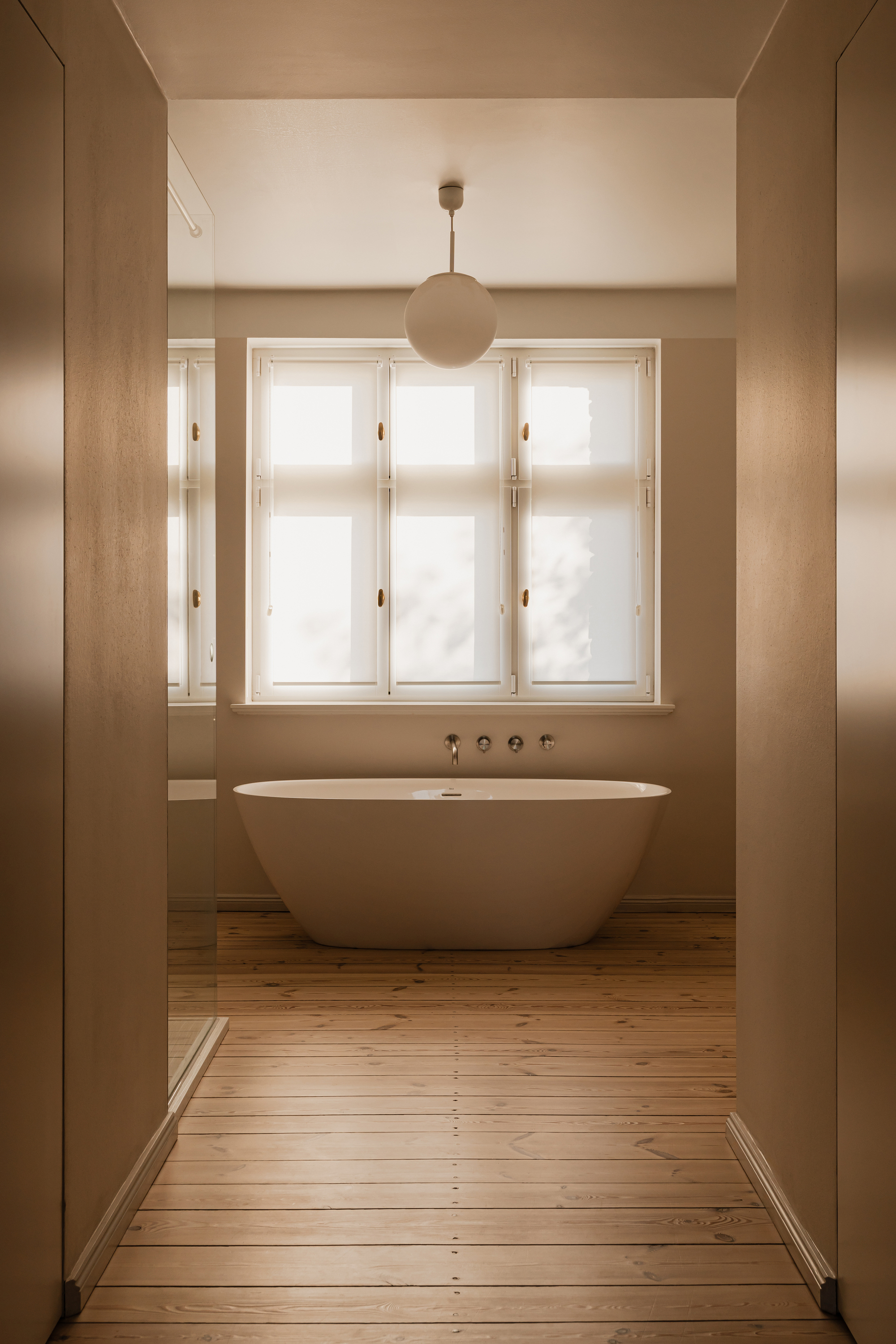 Freestanding white bathtub beneath a frosted casement window, seen between two wall planes in diffused daylight