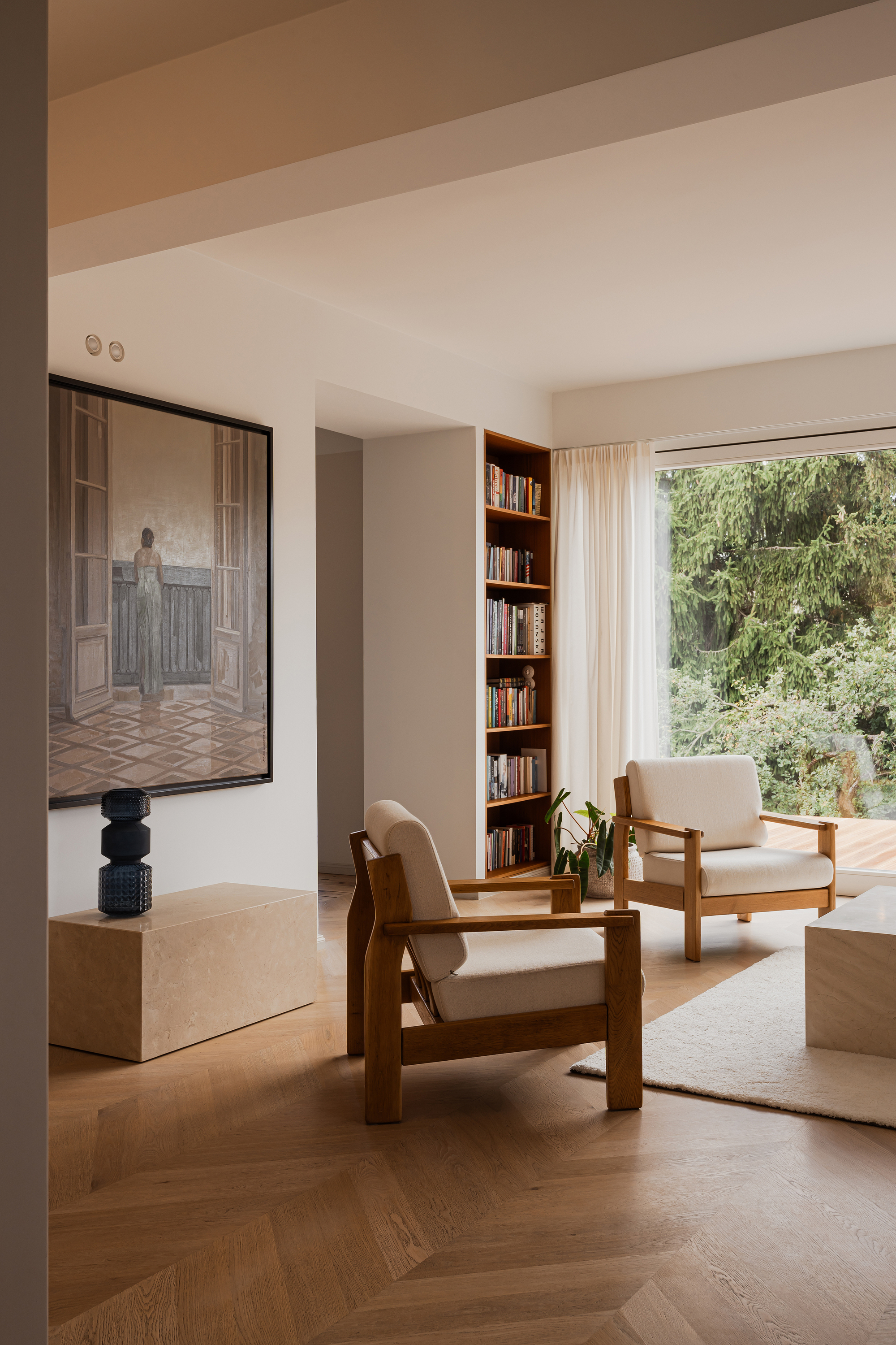 Timber lounge chairs and a travertine block table on herringbone oak, with a tall bookshelf and a garden view through the window
