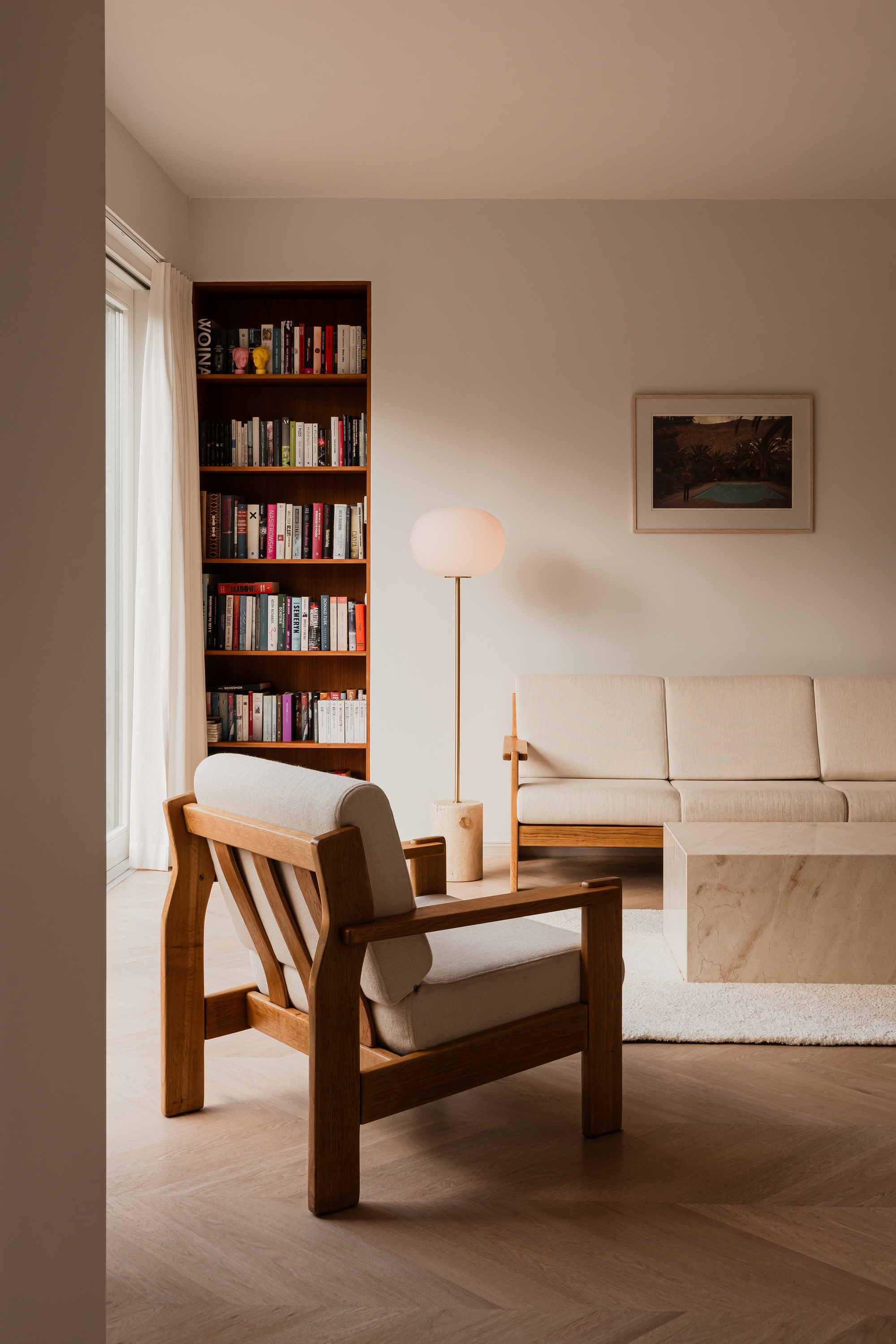 Wooden-framed armchair in the foreground, with a floor lamp, cream sofa and full bookshelf behind in soft overcast light