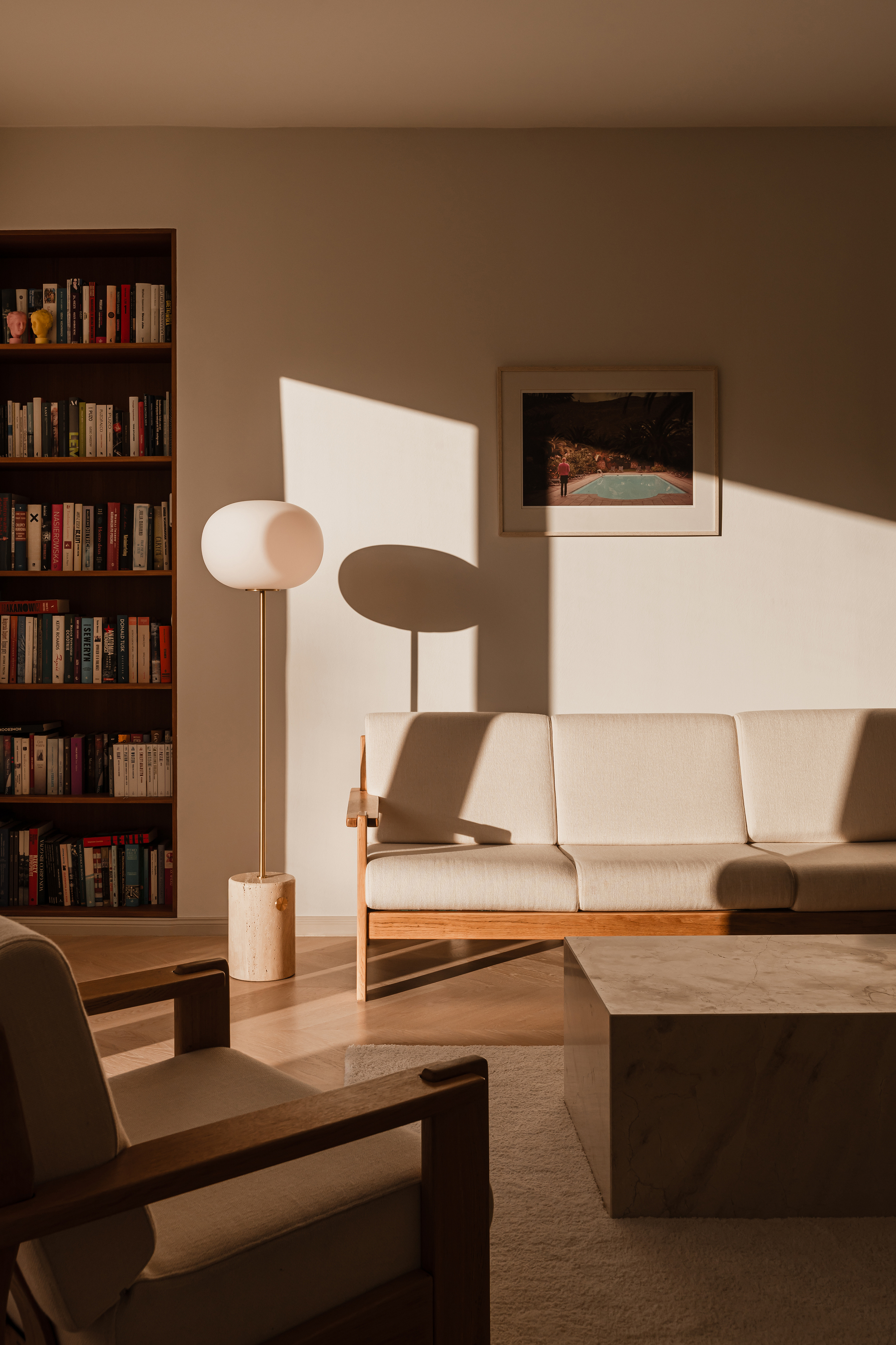 Late sun casts hard shadows across the wall behind the cream sofa, round floor lamp and travertine coffee table