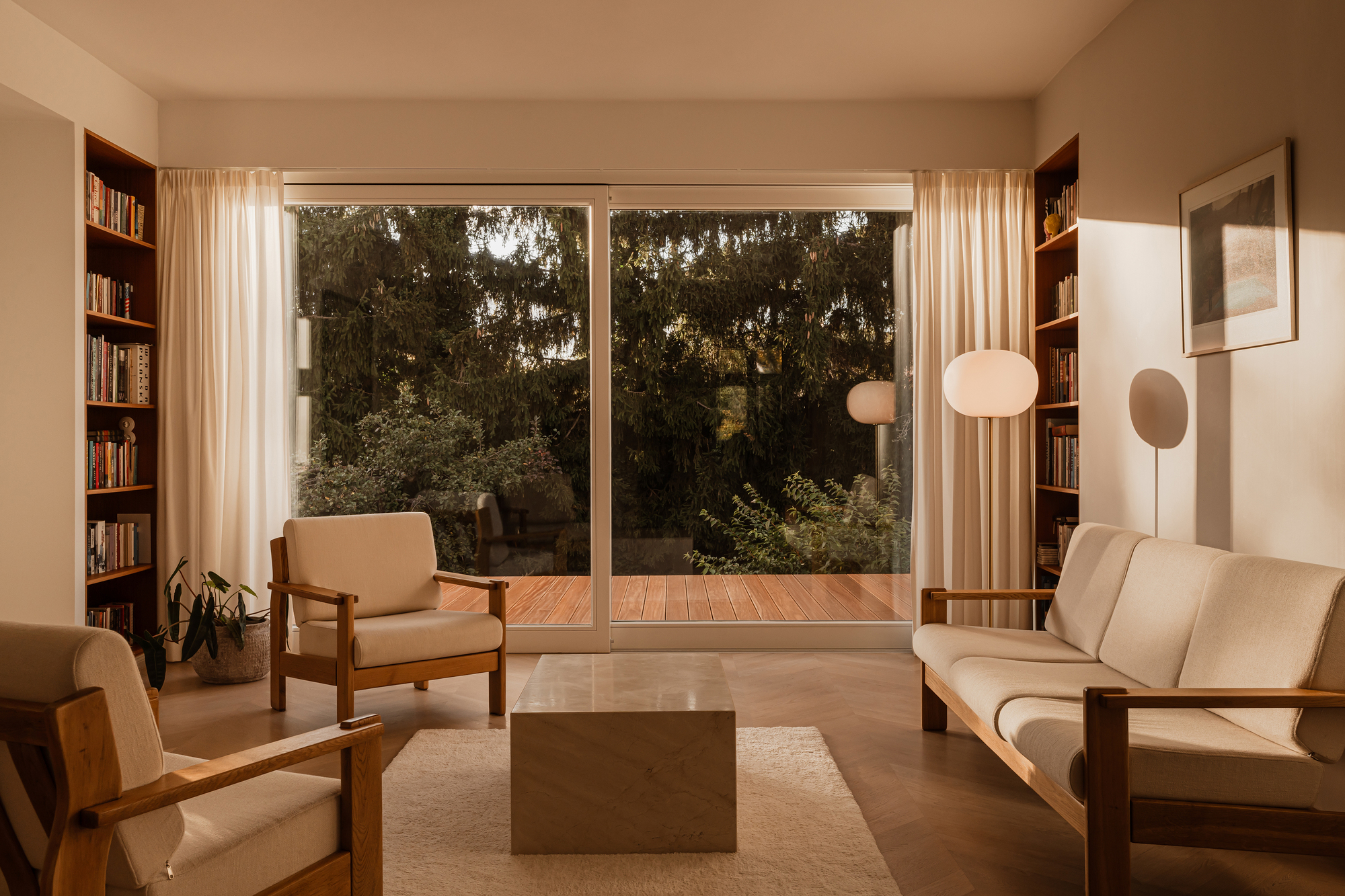 Sitting room at golden hour, with sheer curtains framing a sliding glass door that opens to a timber deck and dark conifers