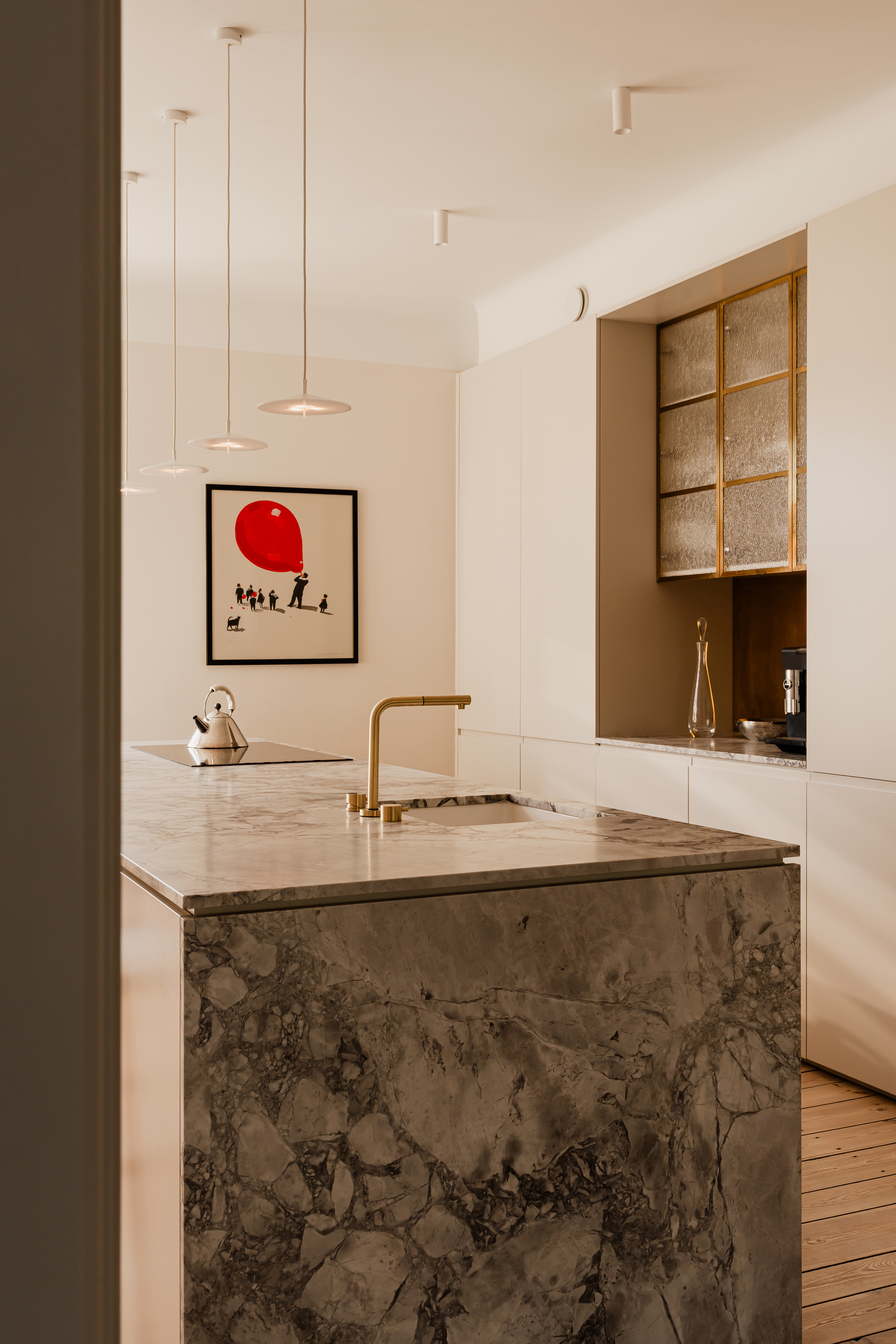 Kitchen island clad in rough grey marble with a brass tap, beneath pendant lamps and a framed red balloon print