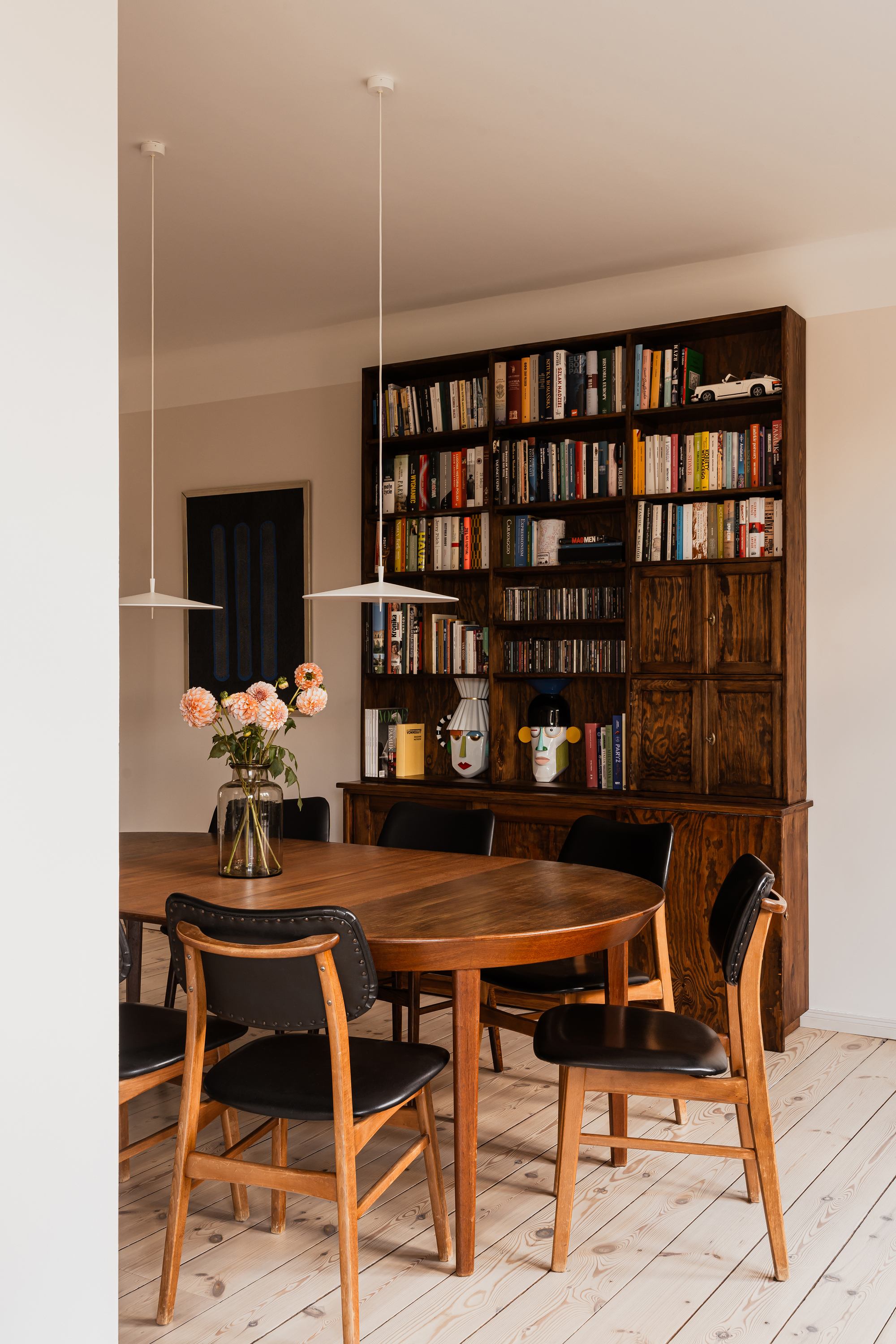 Dark timber bookcase and oval dining table with black leather chairs, pendant lamps and dahlias in a glass vase