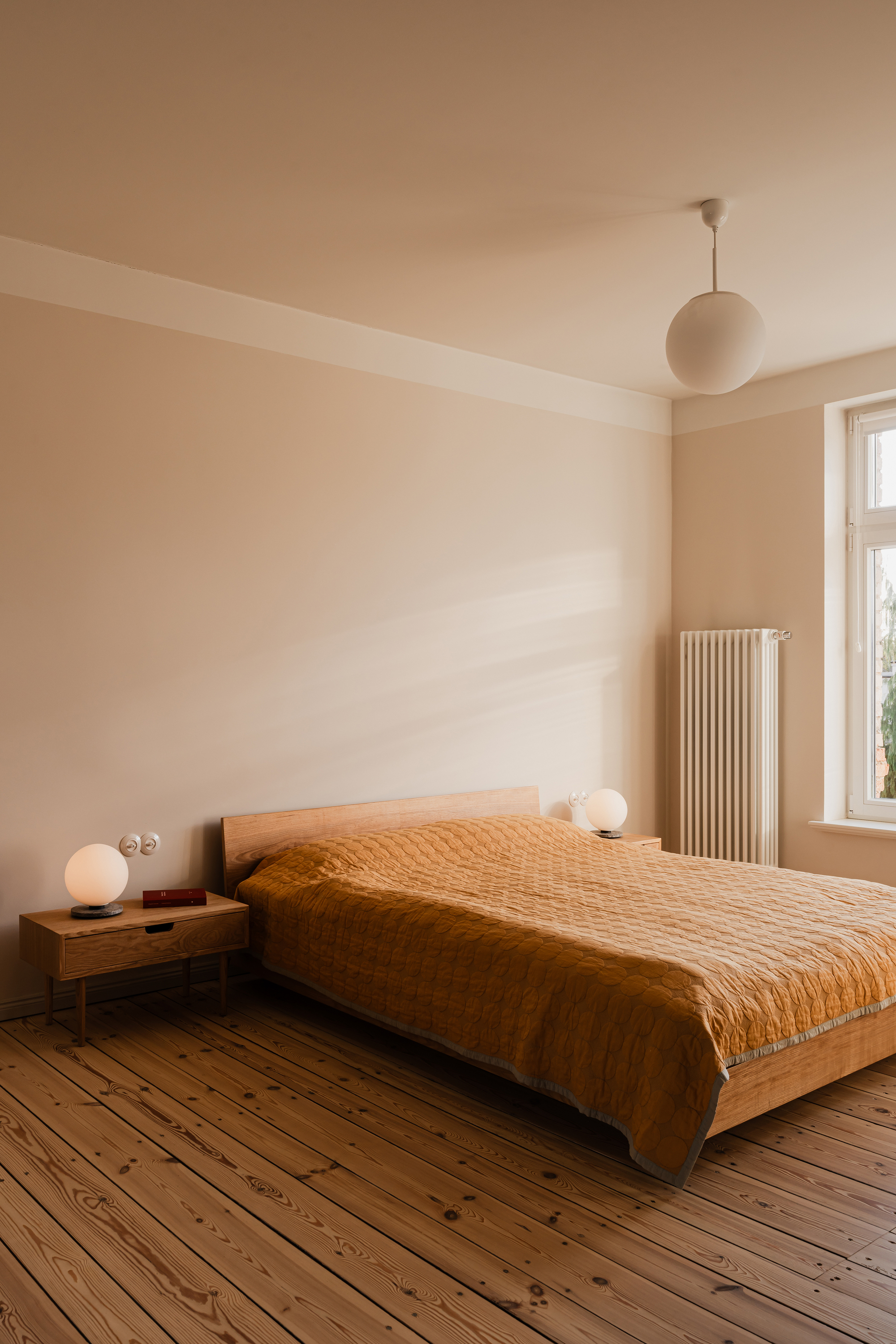 Bedroom with a timber bed and mustard quilt, globe lamps and a radiator beside a window, sunlight on the wall
