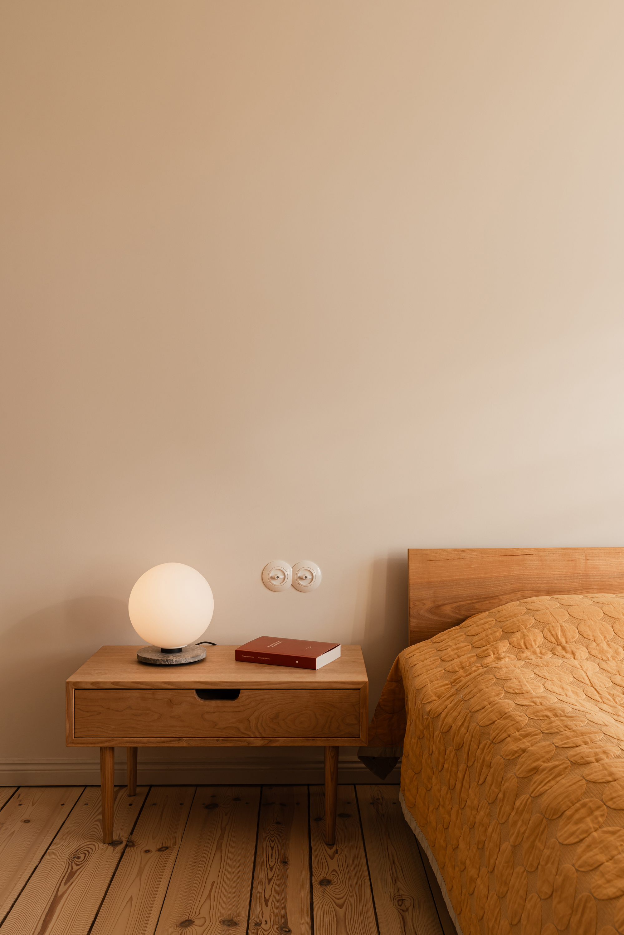 Oak bedside table with a globe lamp and red book, porcelain double switch on the wall above the pine floor
