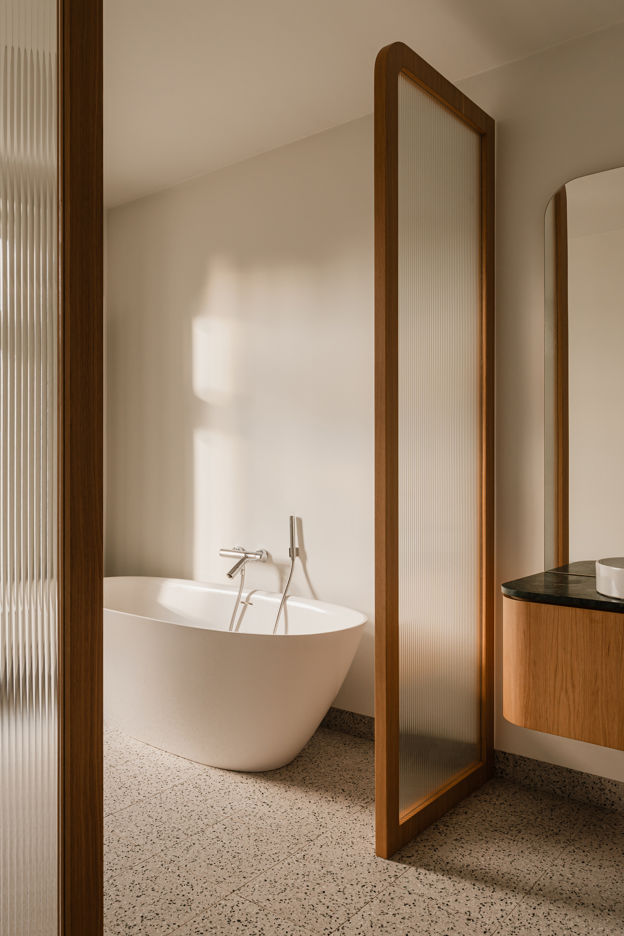 Freestanding white bathtub behind a fluted glass panel in an oak frame, with soft sunlight falling across the terrazzo floor