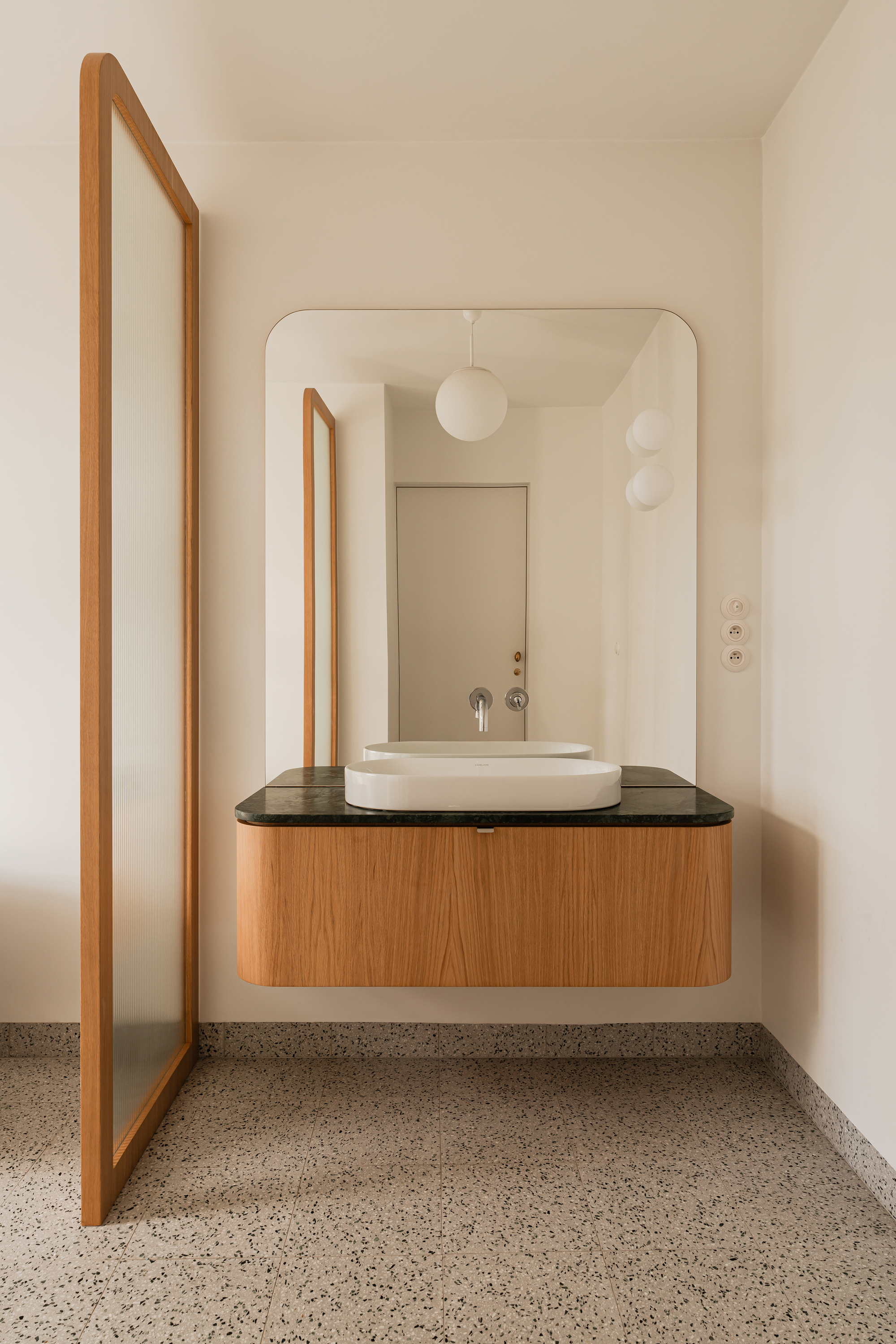 Floating oak vanity with a green stone top and white basin below a rounded mirror, beside a fluted glass screen