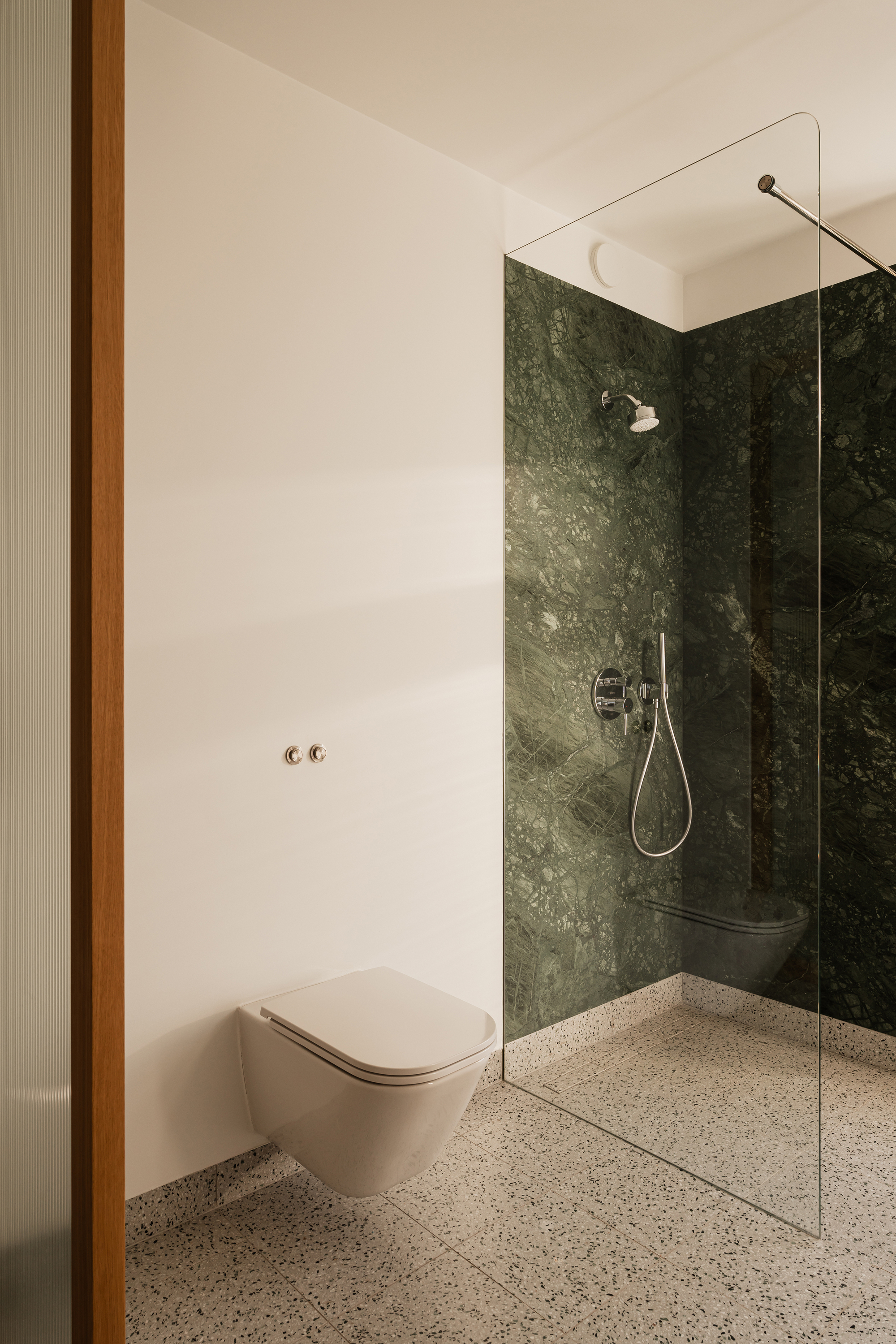 Wall-hung toilet beside a glass-screened shower lined in dark green marble, with afternoon light raking across the plaster wall