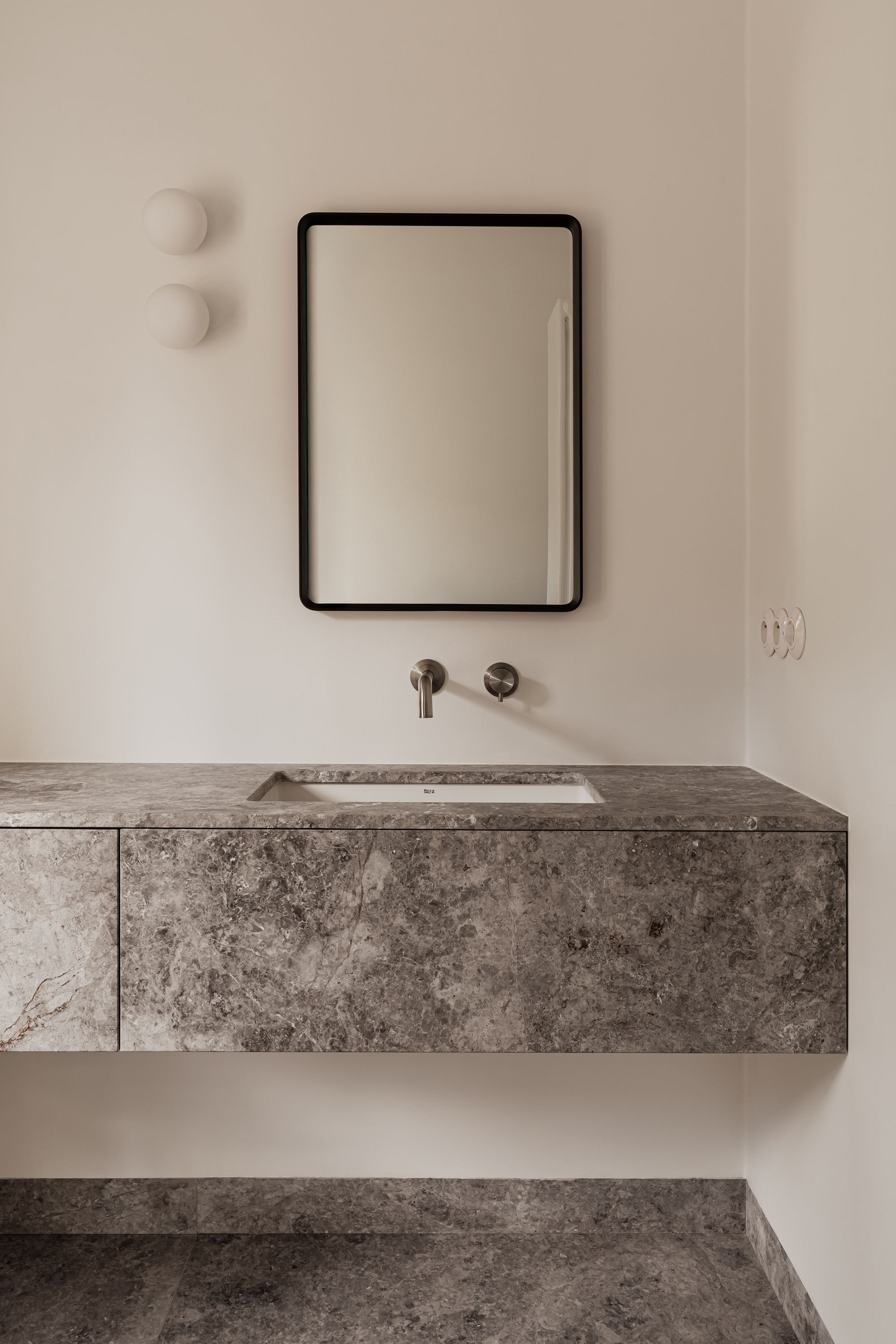 Floating grey stone vanity with an undermount basin, wall-mounted tap and black-framed mirror under two globe lights