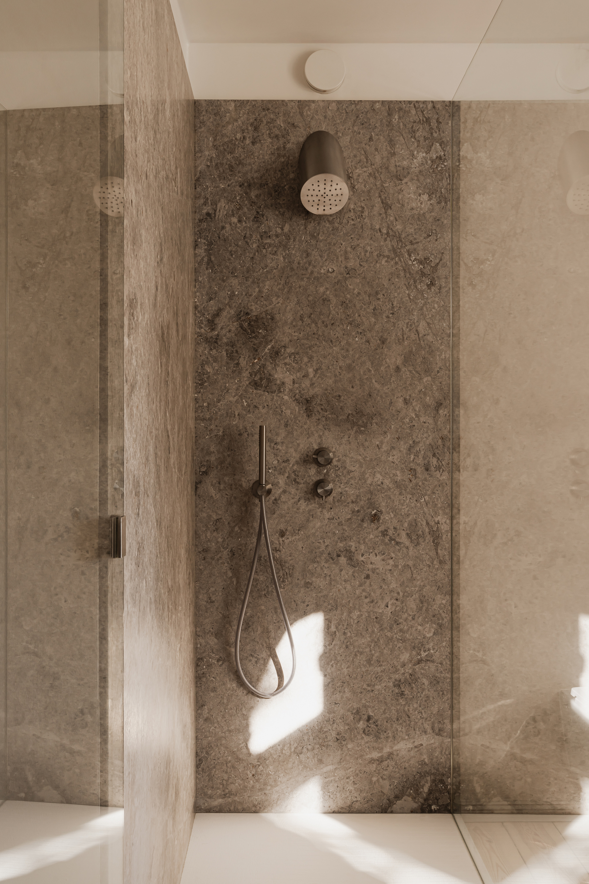 Shower alcove with a brown stone slab wall, cylindrical rain head and hand shower, with sunlight patches on the glass and tray