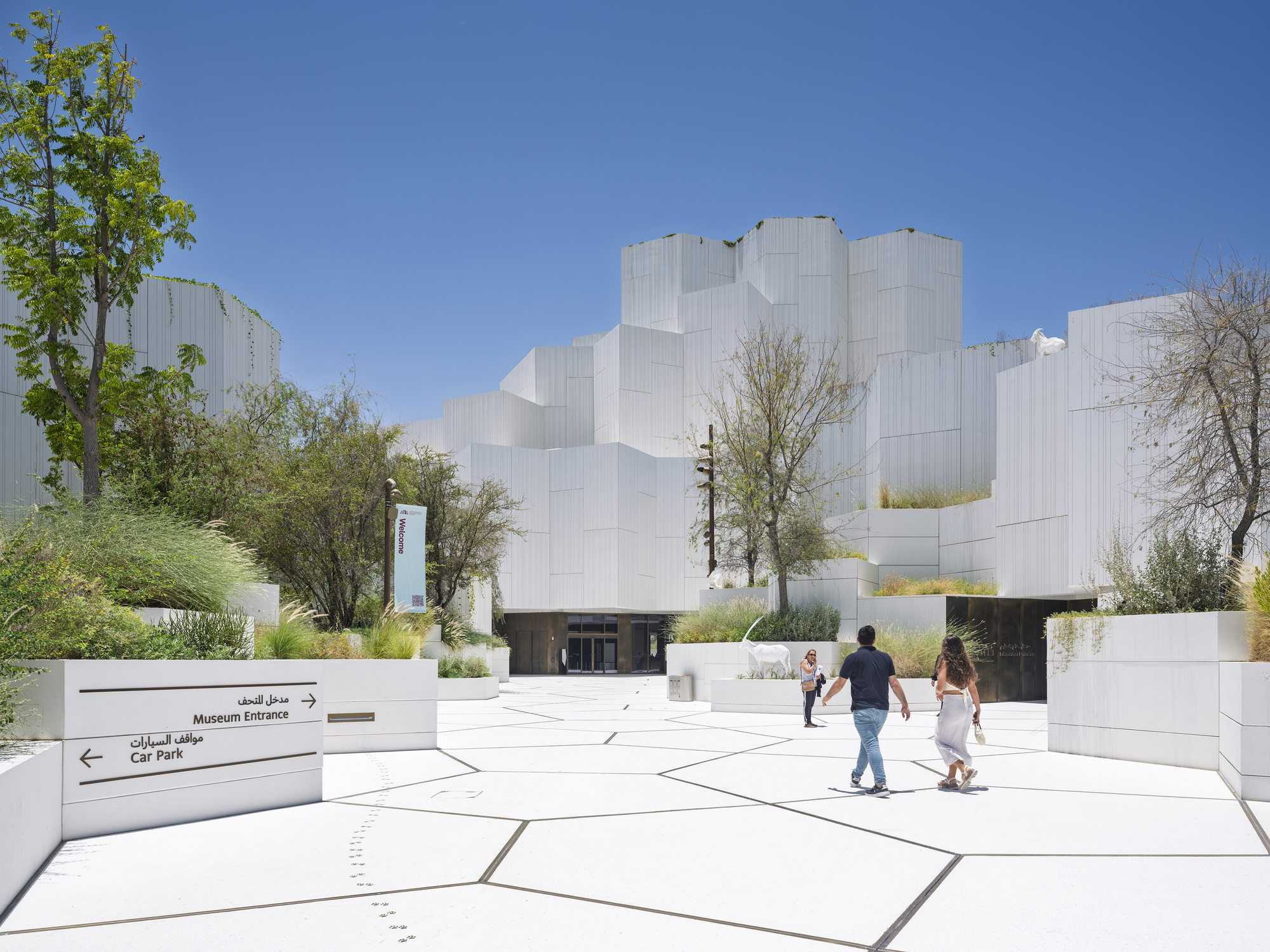 White hexagon-paved plaza with bilingual wayfinding signs and stacked white volumes beyond, two visitors walking under a clear blue sky