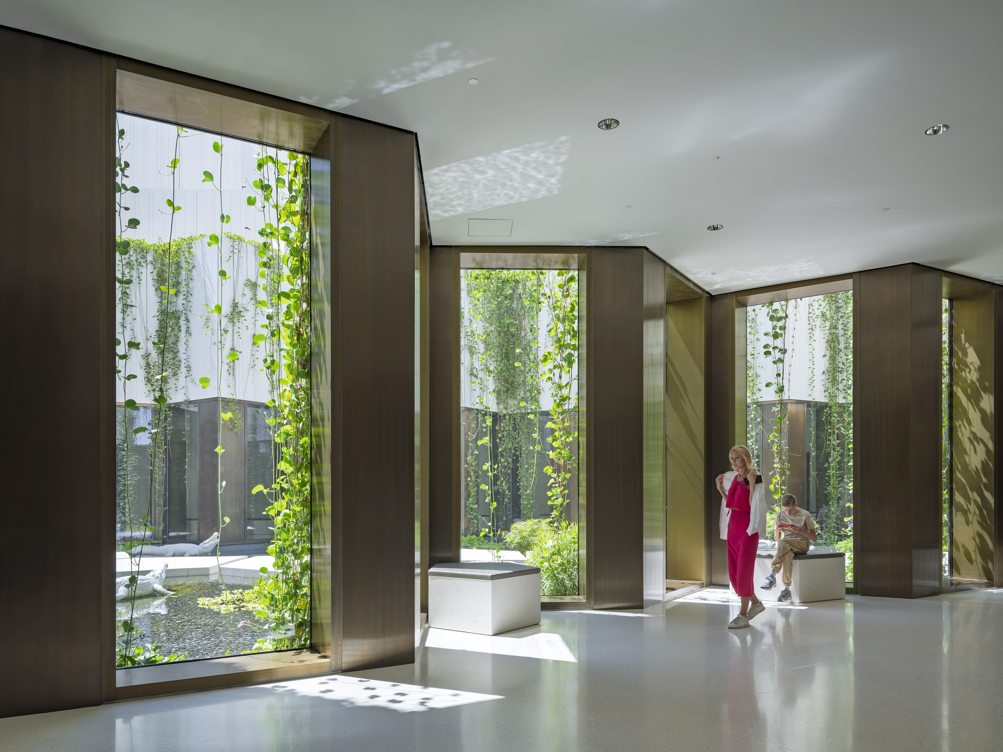 Bronze-clad window bays framing hanging vines and a courtyard pool, with a woman in pink standing in dappled sunlight