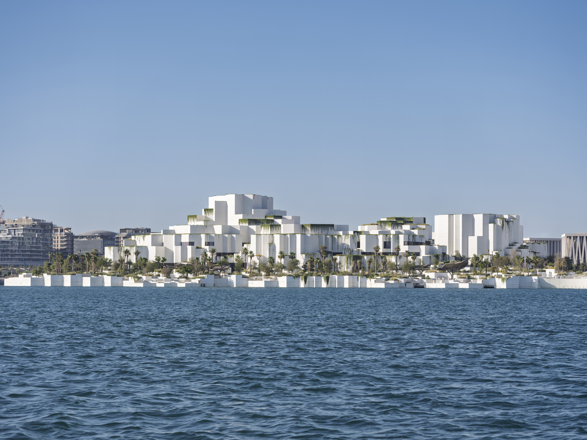 Cluster of stepped white blocks with rooftop greenery seen across open blue water on a hazy clear day