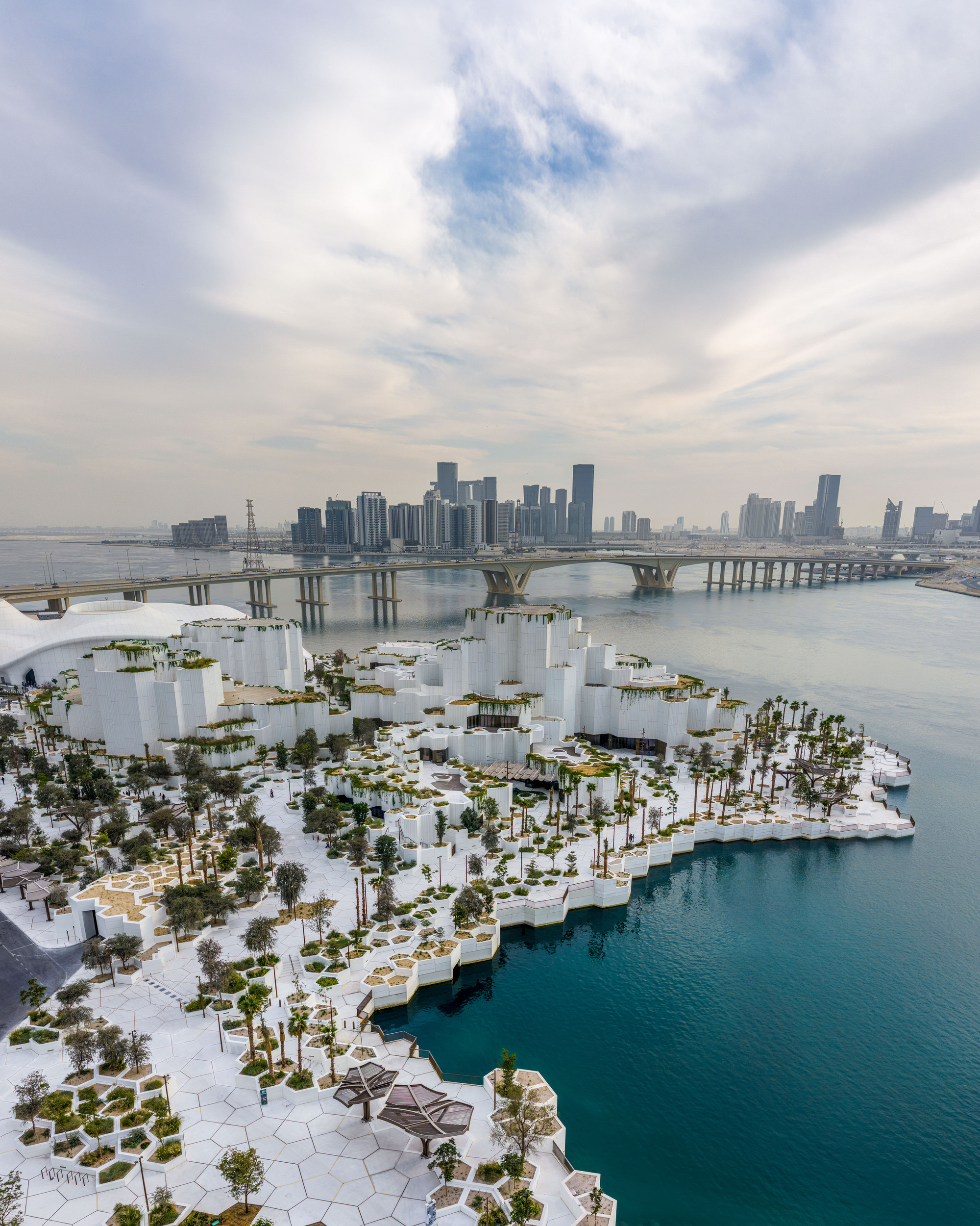 Aerial view of white stepped volumes and hexagon-paved terraces with palms on a waterfront peninsula under a cloudy sky