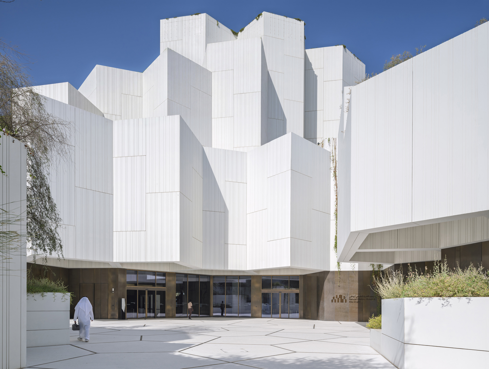 Entrance plaza facing faceted white ribbed volumes above a glazed base, with a figure in white robes crossing the paving