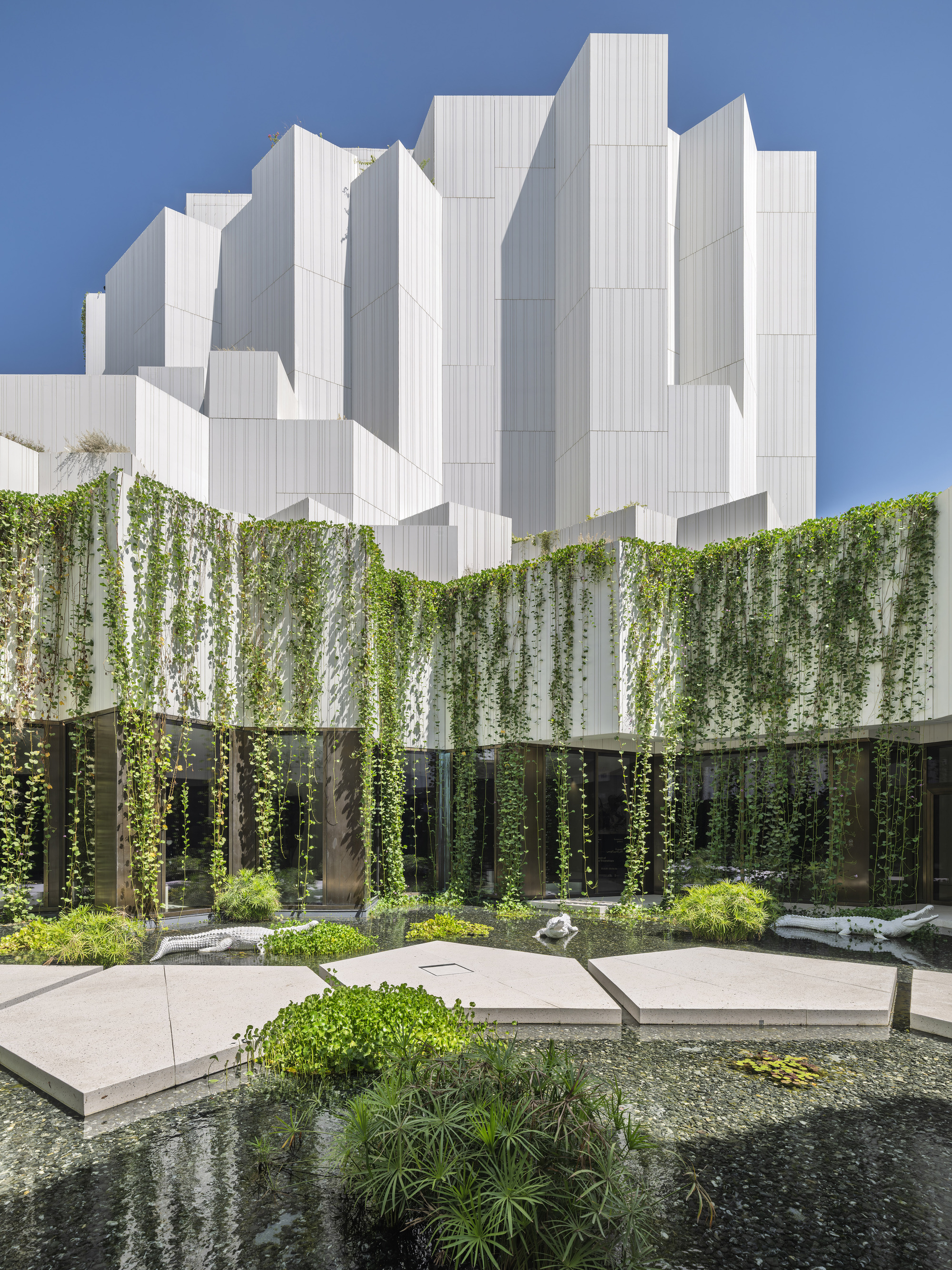 Pool courtyard with angular stone stepping platforms and vines hanging from a white facade beneath stacked white towers in clear sun