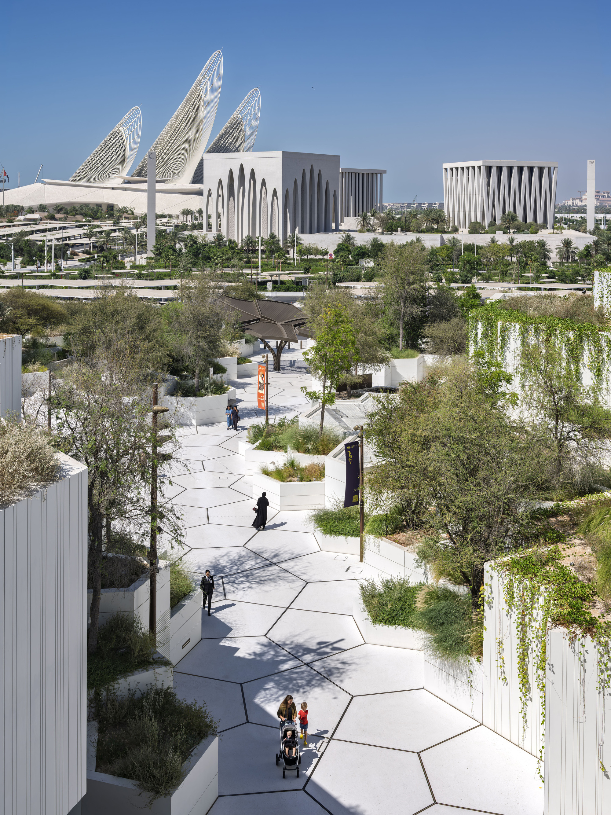 Hexagonal paved rooftop path between planted white beds, with a family and pedestrians walking toward distant white institutional buildings under clear midday s