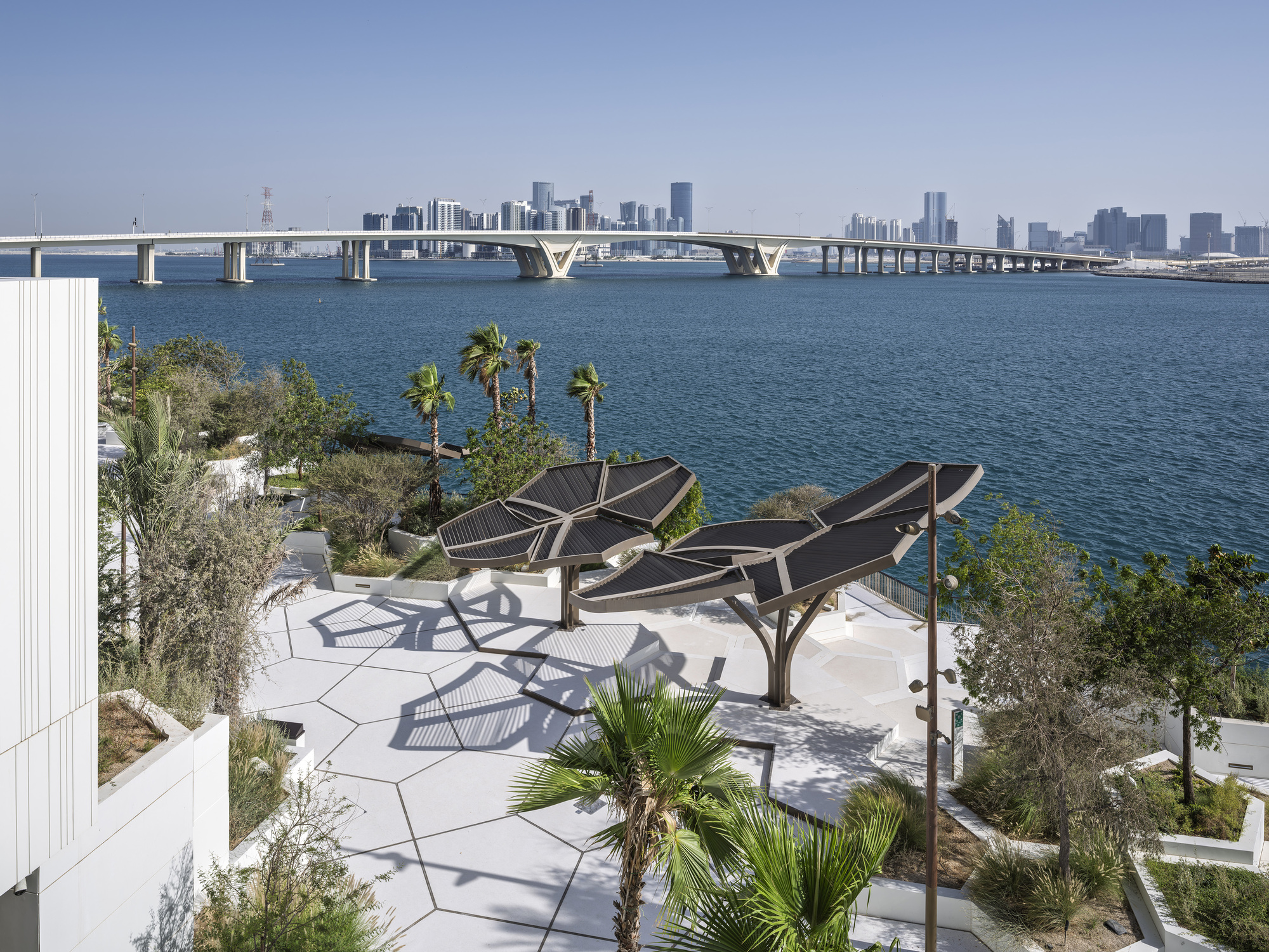 Rooftop terrace with leaf-shaped solar canopies and palms, overlooking a bridge and distant skyline across blue water in hazy daylight