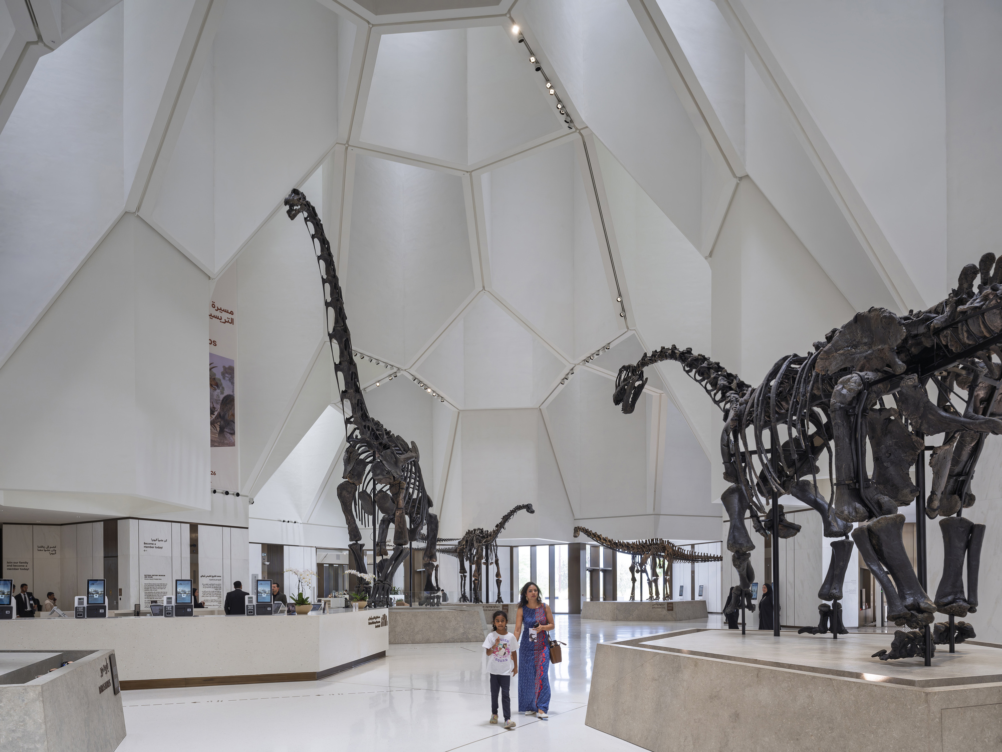 Atrium lobby with faceted white ceiling and mounted dinosaur skeletons, a woman and child walking across the polished floor
