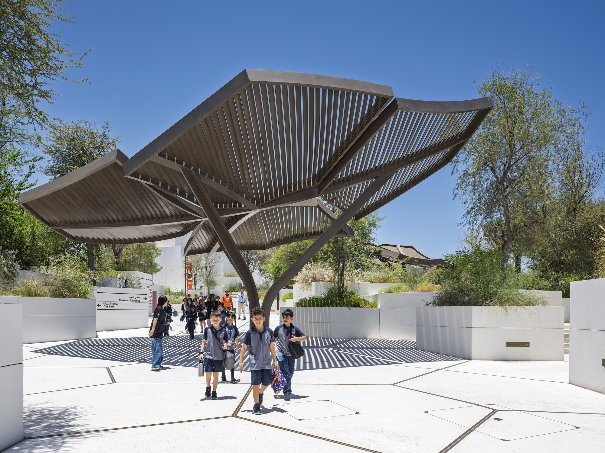 Louvred canopy shading a paved plaza with planters as schoolchildren walk through under a clear blue midday sky